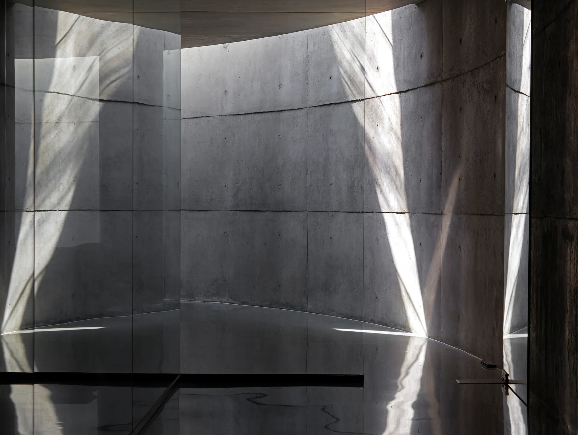 A minimalist interior space features curved raw concrete walls and a polished dark reflective floor illuminated by beams of natural light.