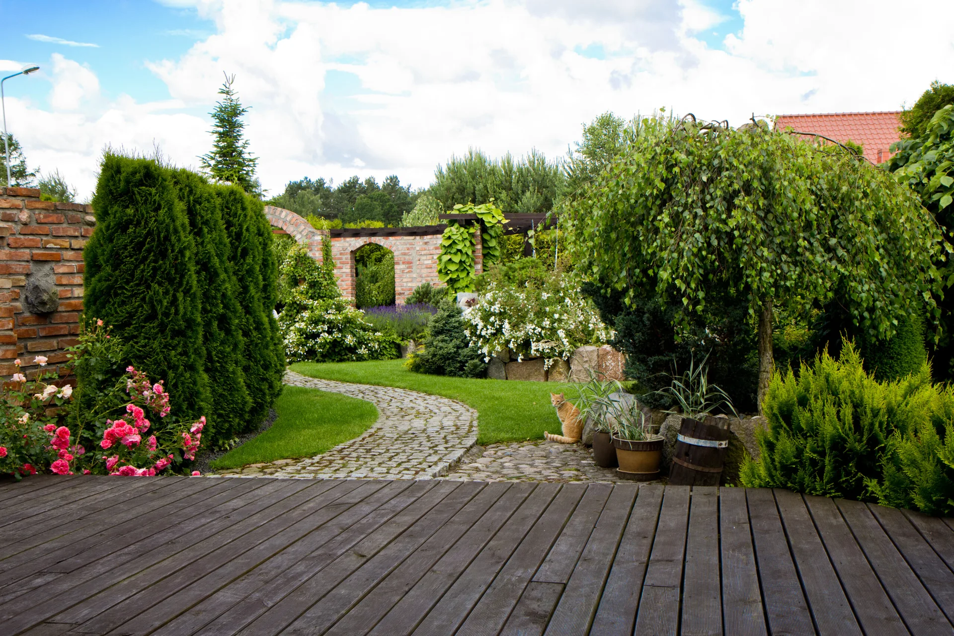 A dark wooden deck overlooks a landscaped backyard garden with a winding cobblestone path, green hedges, pink rose bushes, and brick archways.
