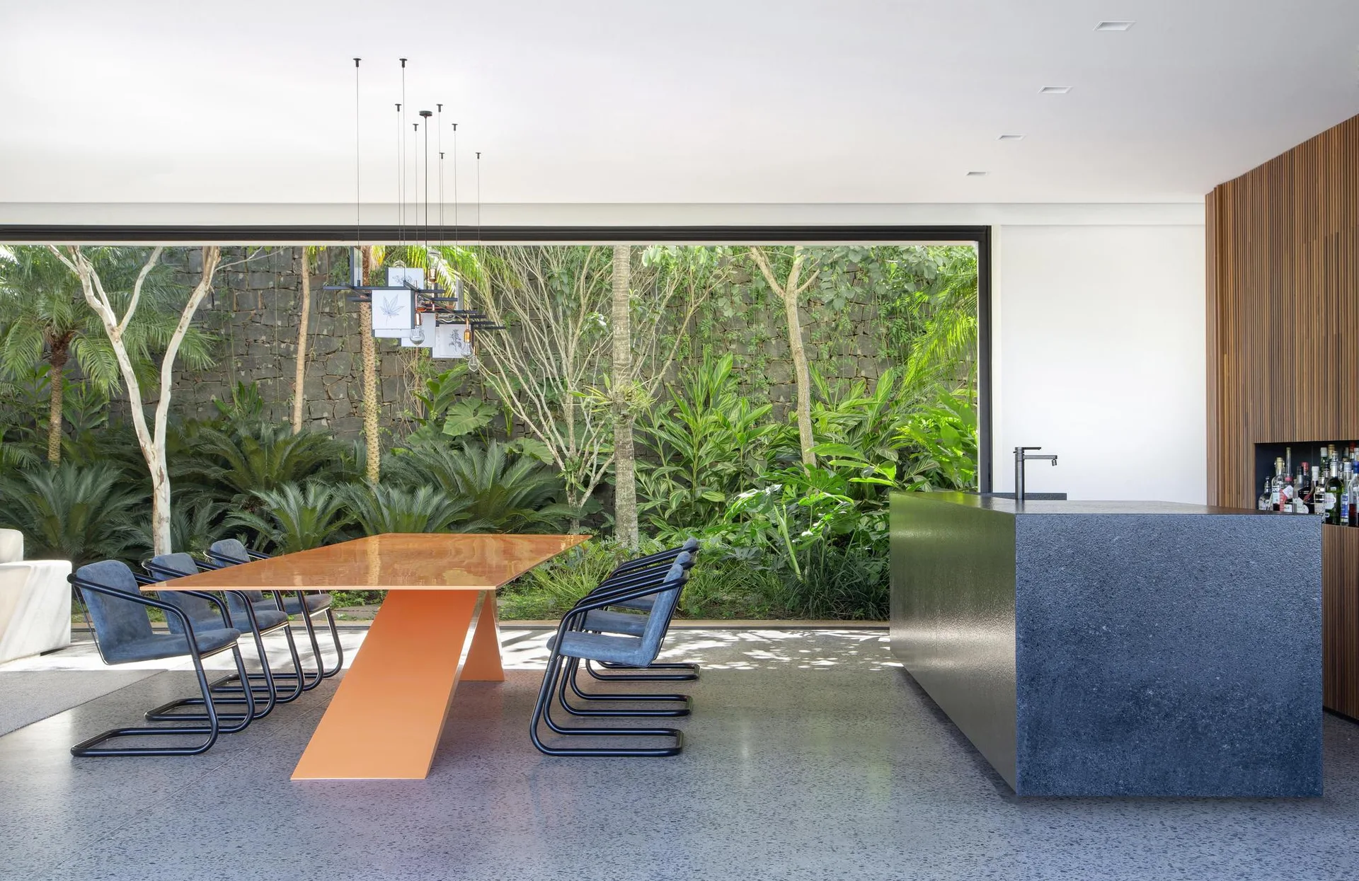 A modern dining room features a long orange table and blue chairs beside a large glass window looking out at a lush garden.