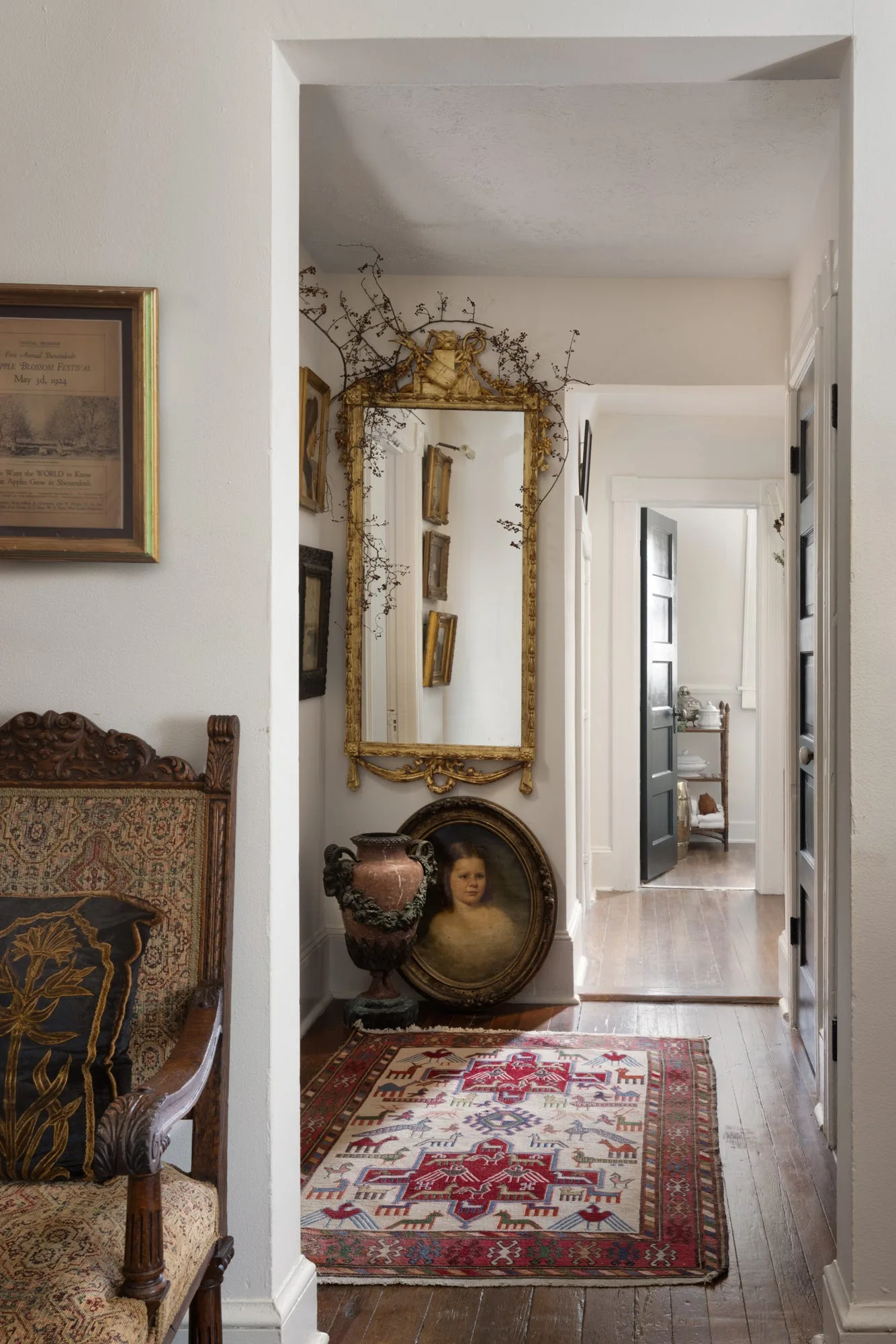 A traditional hallway featuring a patterned red rug, an ornate gold mirror, a stone urn, and an oval portrait painting on dark wood floors.