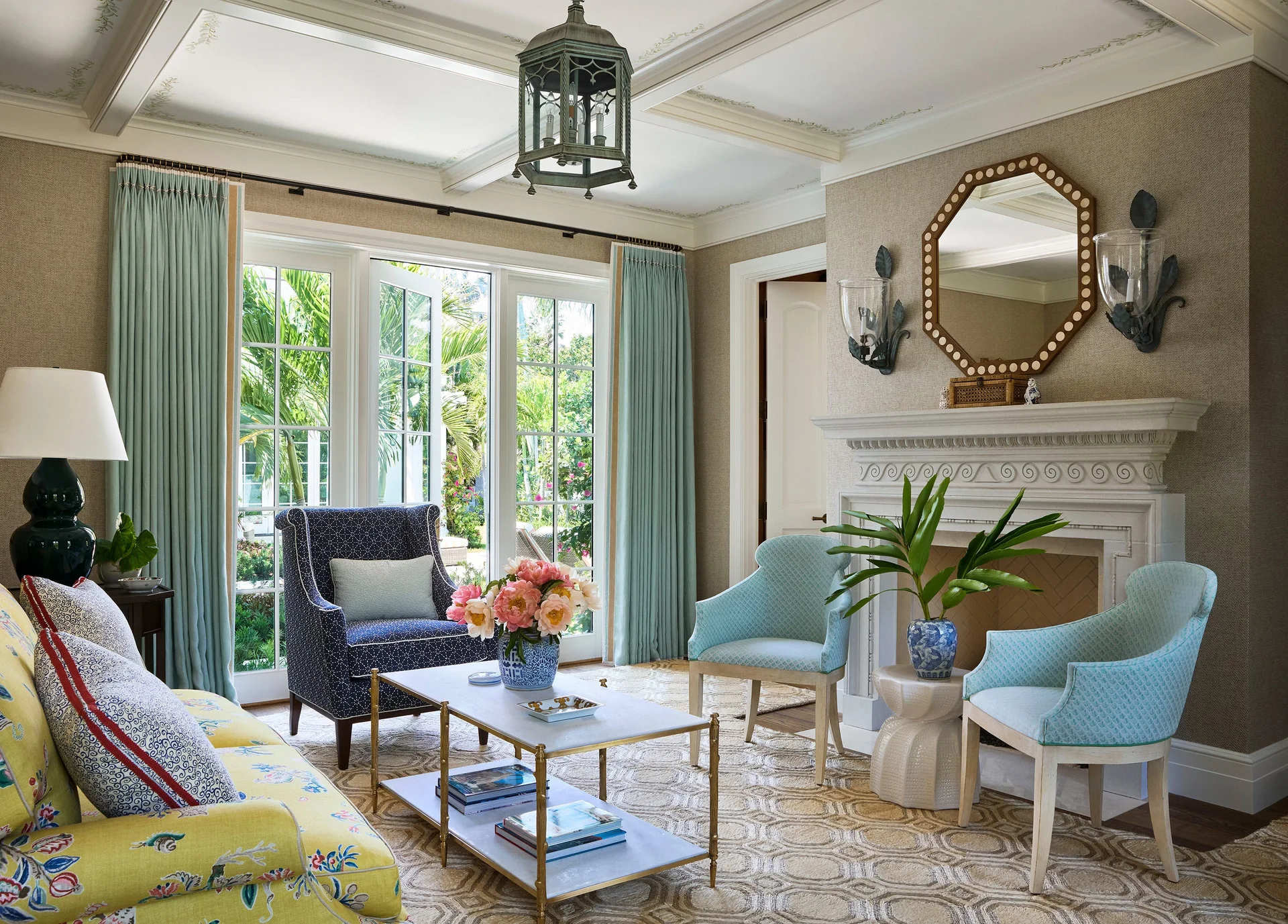 A bright living room features a yellow floral sofa, a pair of blue armchairs by a white fireplace, and glass doors looking out to a garden.