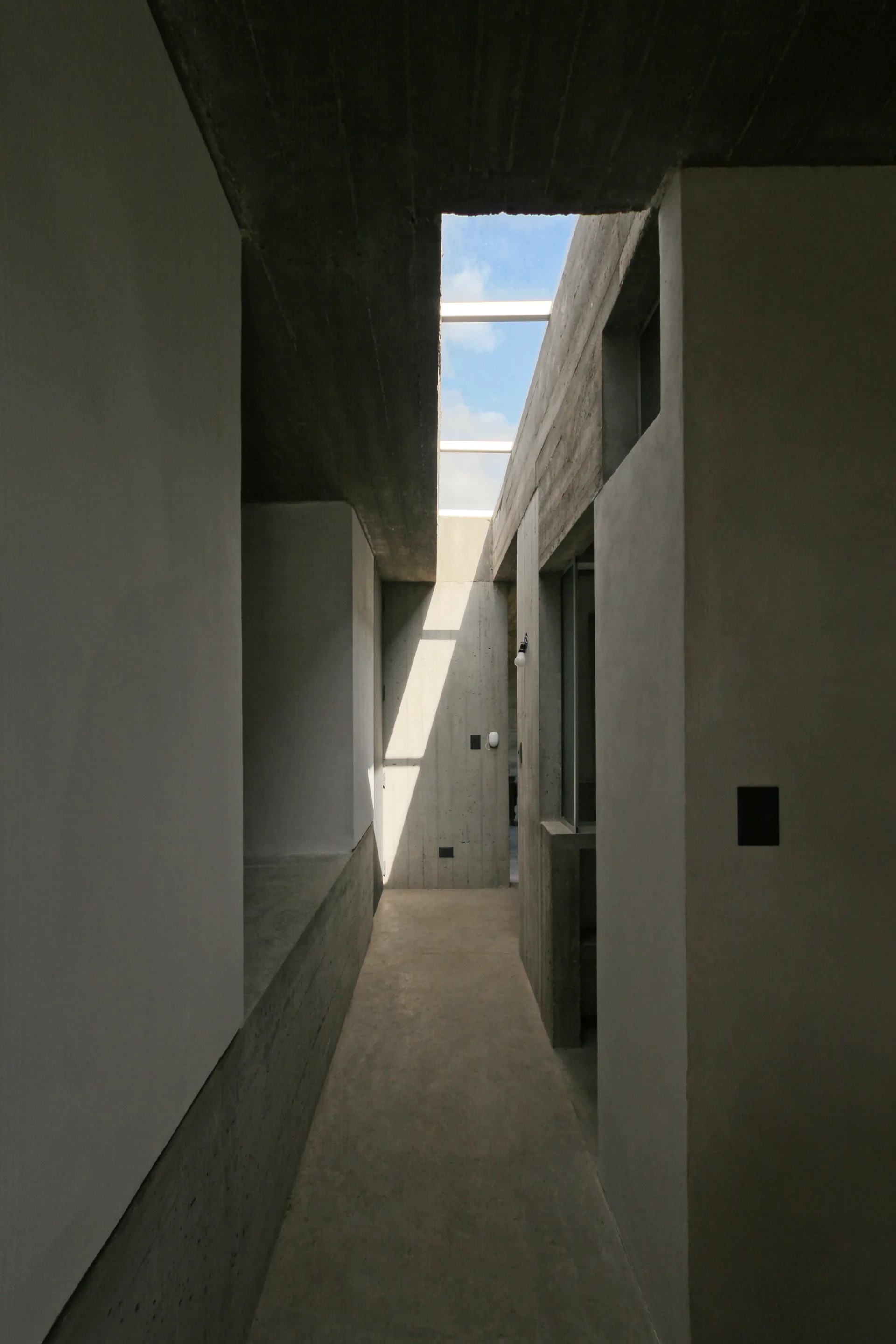 A narrow gray concrete corridor features matte cement walls, a matching concrete floor, and a linear skylight opening to the blue sky.