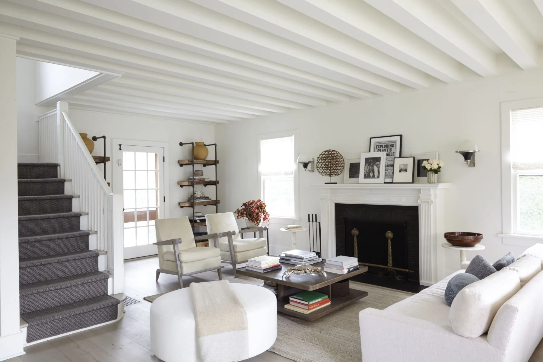 A bright white living room featuring a white beamed ceiling, a central fireplace with decorated mantel, and a dark carpeted staircase.