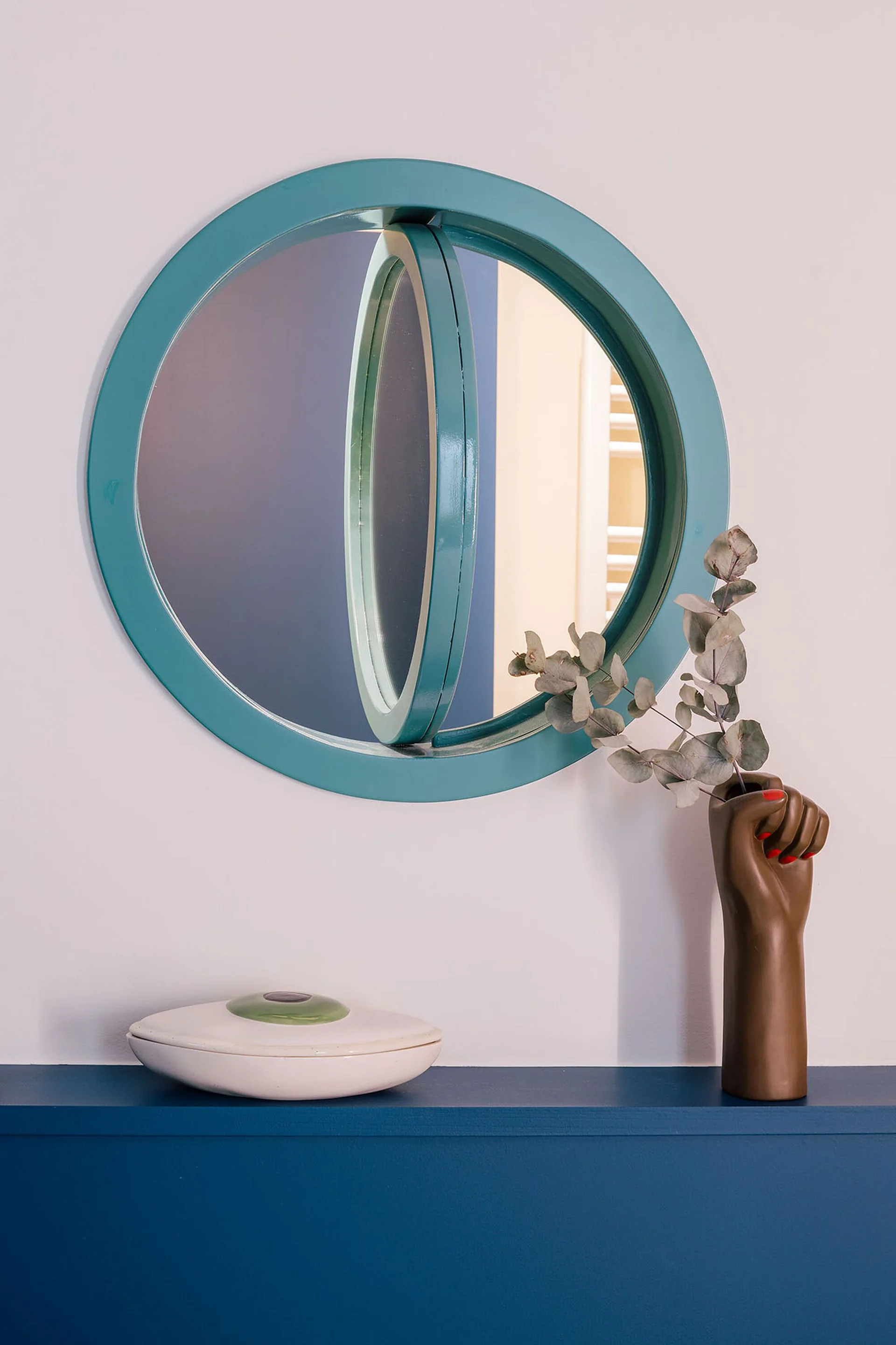 A circular teal pivot mirror hangs on a white wall above a blue shelf holding a fist-shaped ceramic vase and a lidded bowl.