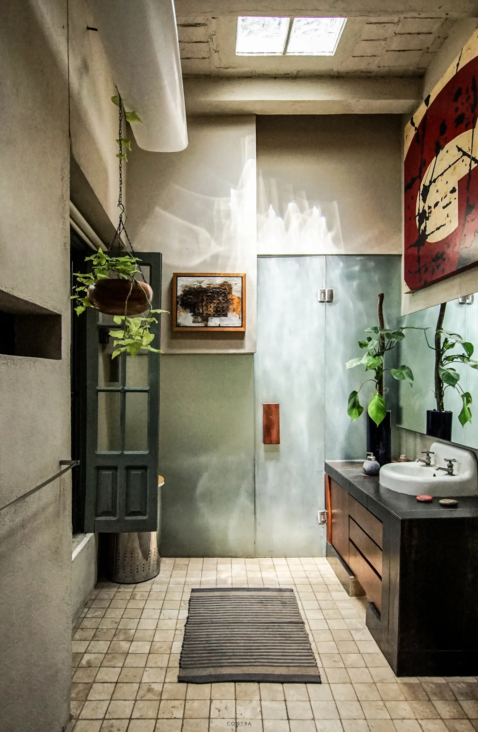 A rustic bathroom features a dark wood vanity with a white sink, textured plaster walls, a frosted glass shower door, and a ceiling skylight.