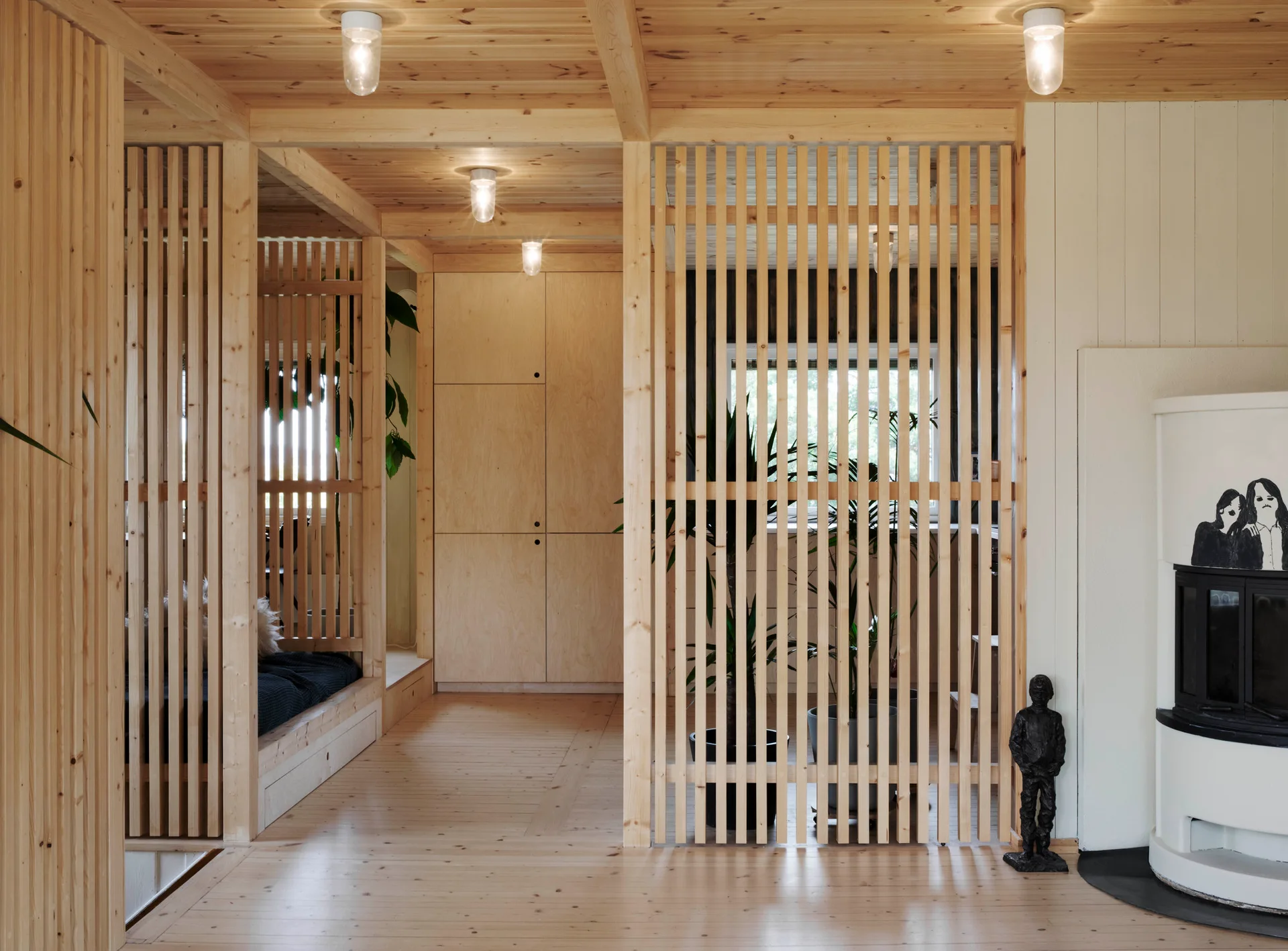 A light wood room features vertical slat room dividers, built-in plywood cabinets, a white tile fireplace, and simple flush-mount ceiling lights.