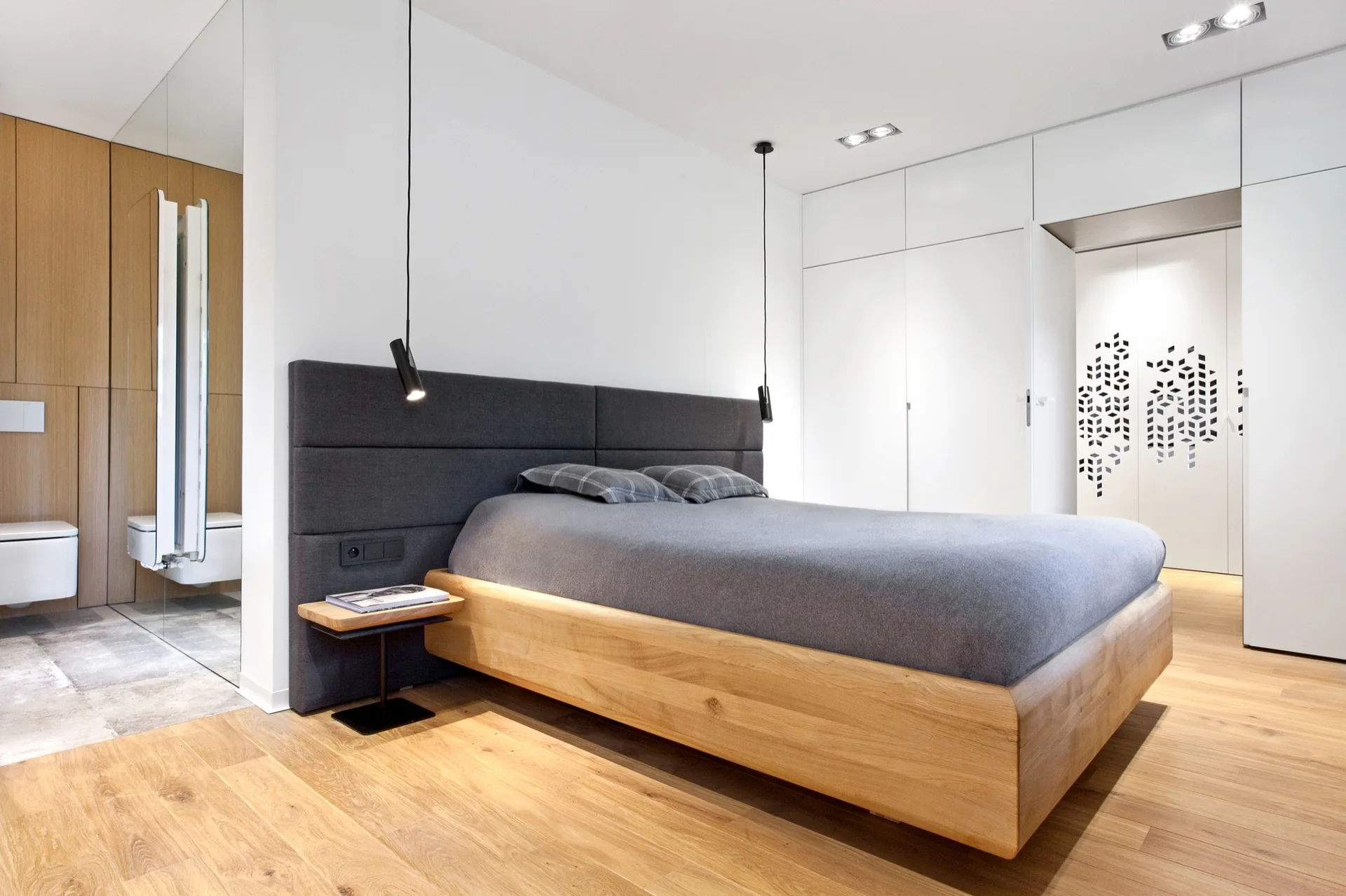 A modern bedroom features a wood-framed bed with a grey upholstered headboard, black hanging pendant lights, and floor-to-ceiling white cabinets.