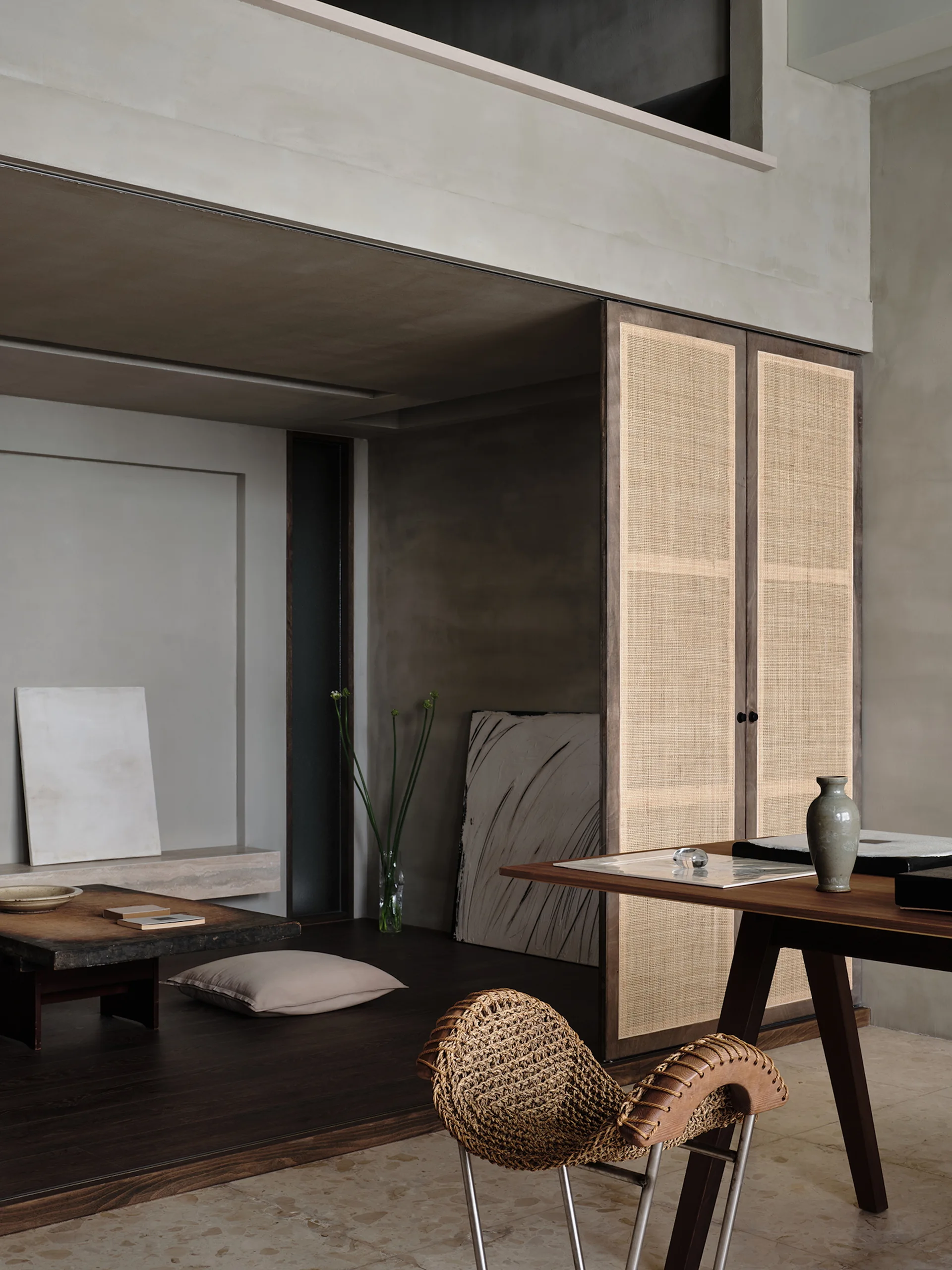 A minimalist room featuring a raised dark wood platform with a low table, plaster walls, a woven metal-framed chair, and tall cane-paneled cabinet doors.