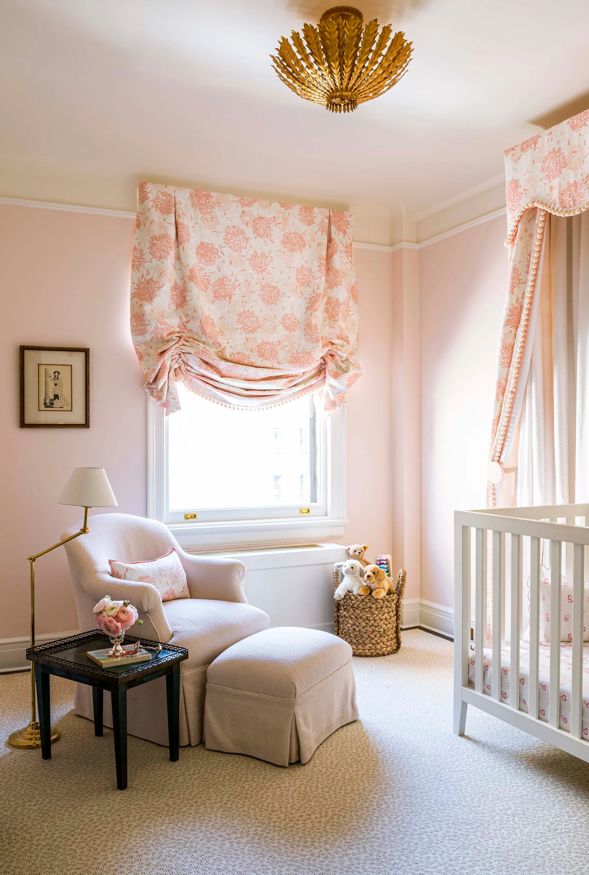 A soft pink nursery features a white wooden crib with floral canopy fabric, a matching armchair with an ottoman, and a gold leaf ceiling fixture.