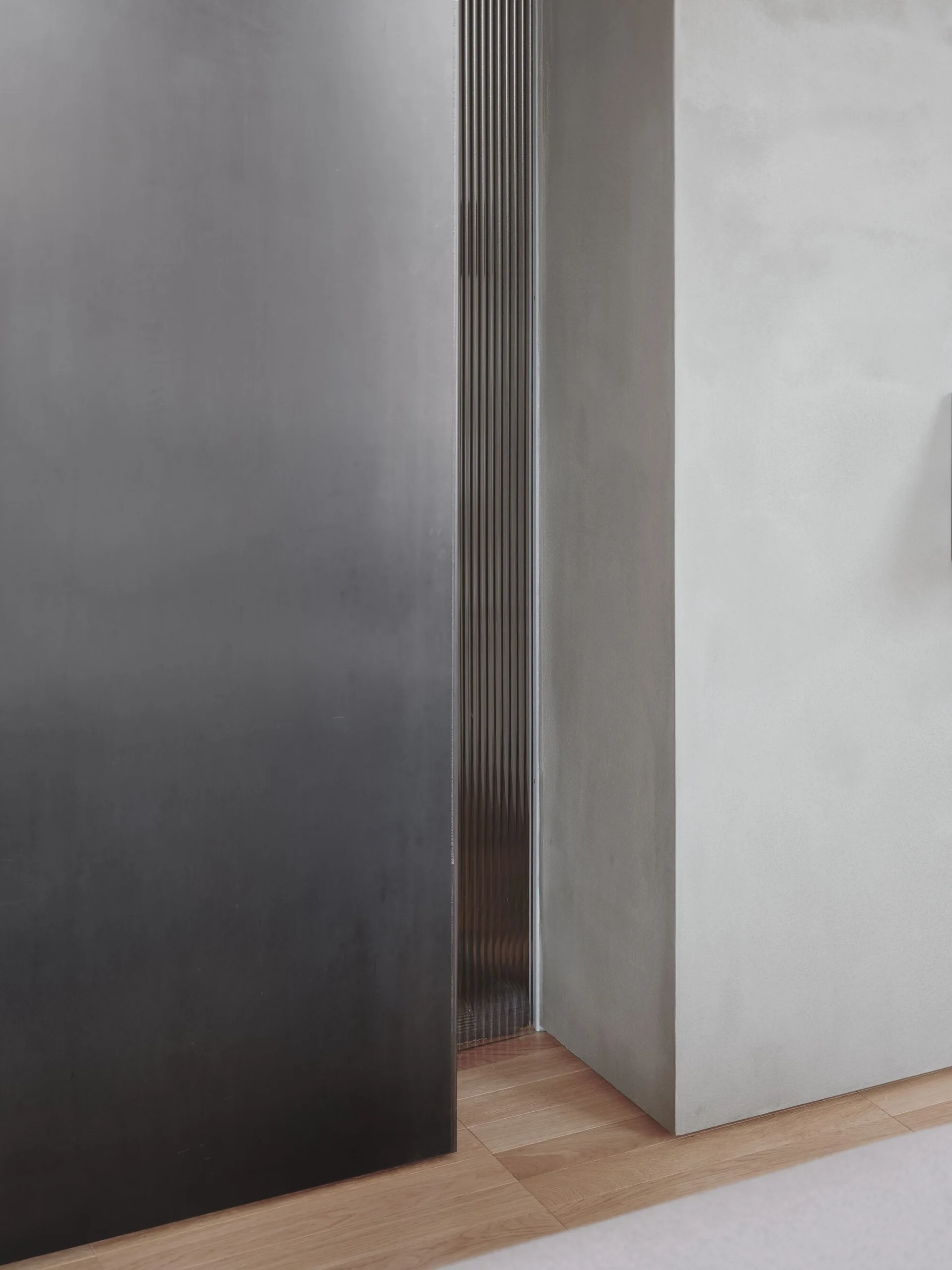 A close-up view of a dark charcoal metal panel next to a narrow vertical fluted metal inlay and a smooth light grey concrete wall.
