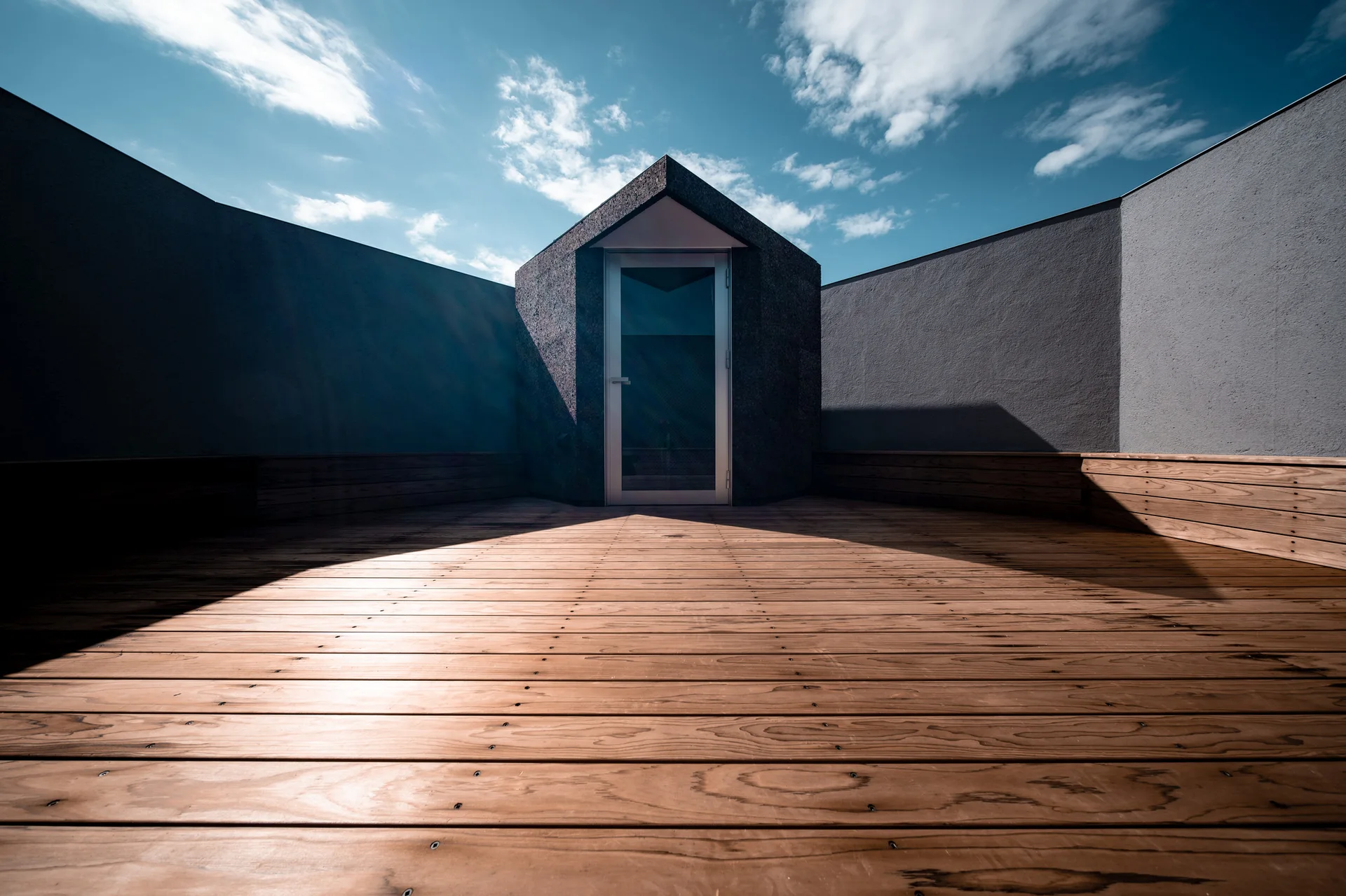 An outdoor rooftop deck features wide wooden planks, a central dark geometric entryway with a glass door, and tall gray privacy walls under a blue sky.