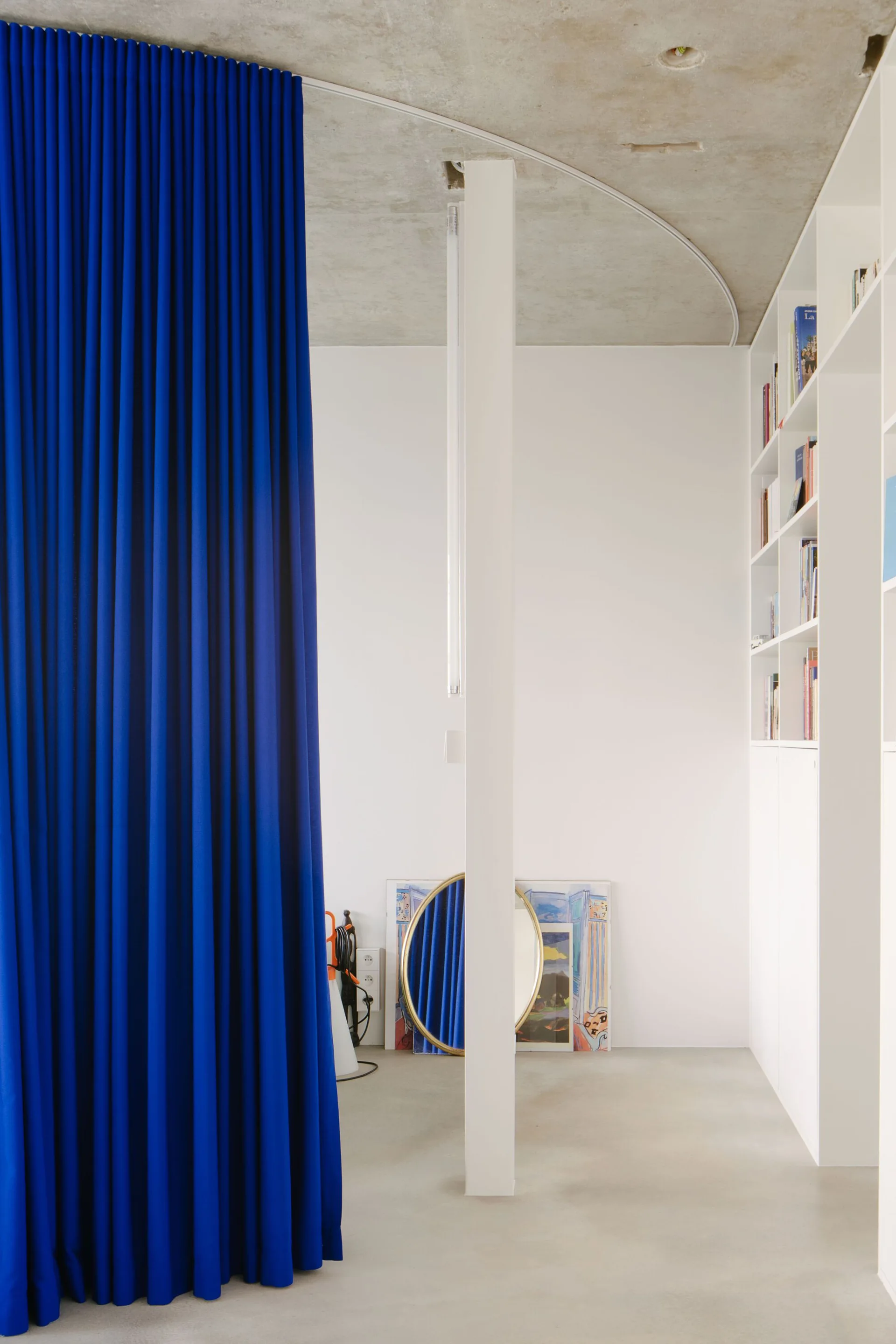 A bright blue floor-to-ceiling curtain hangs on a curved track against a raw concrete ceiling, next to a white structural column and built-in shelving.