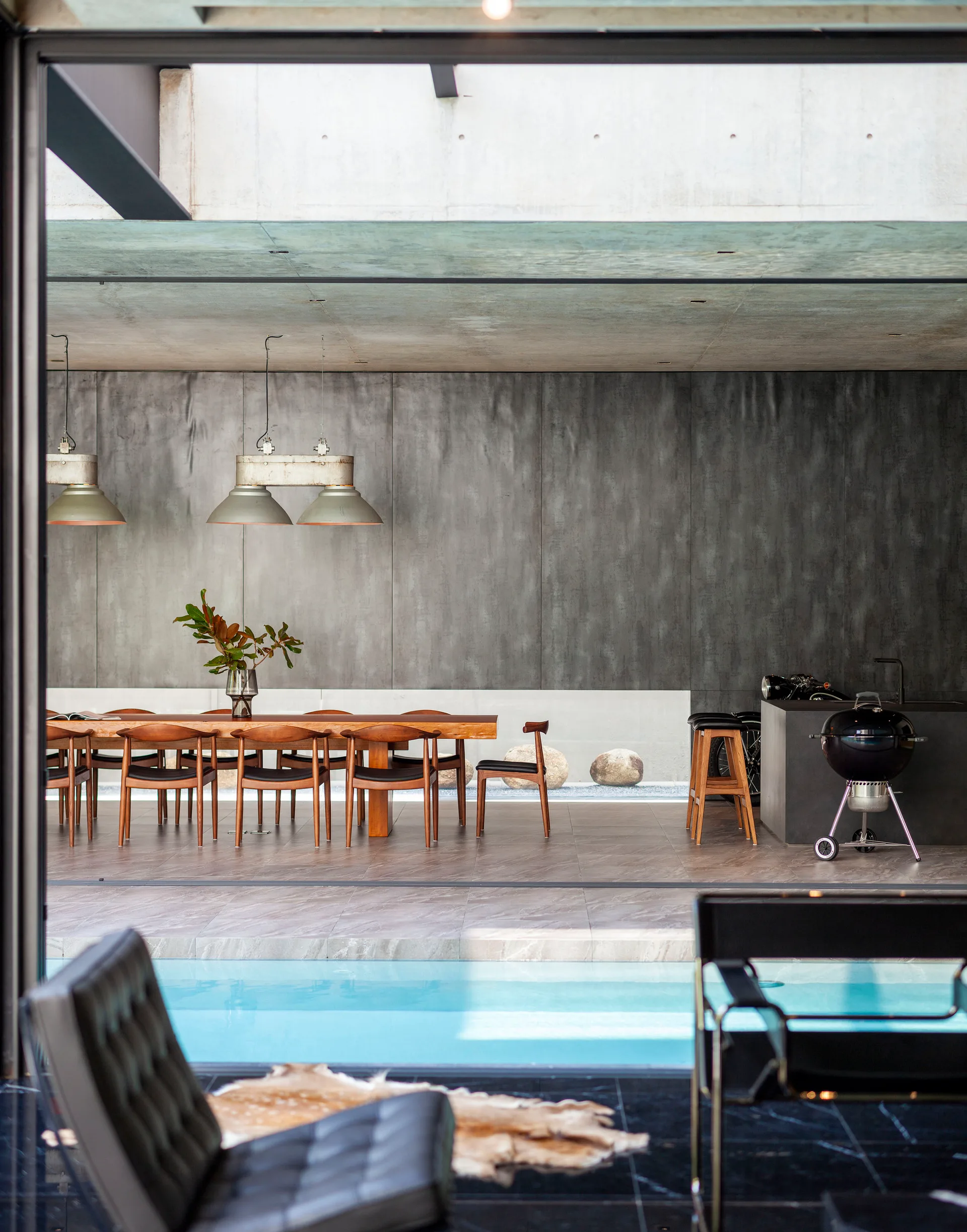 A modern dining area features a long wood table with mid-century chairs, industrial pendant lights, and a smooth concrete wall next to an indoor pool.