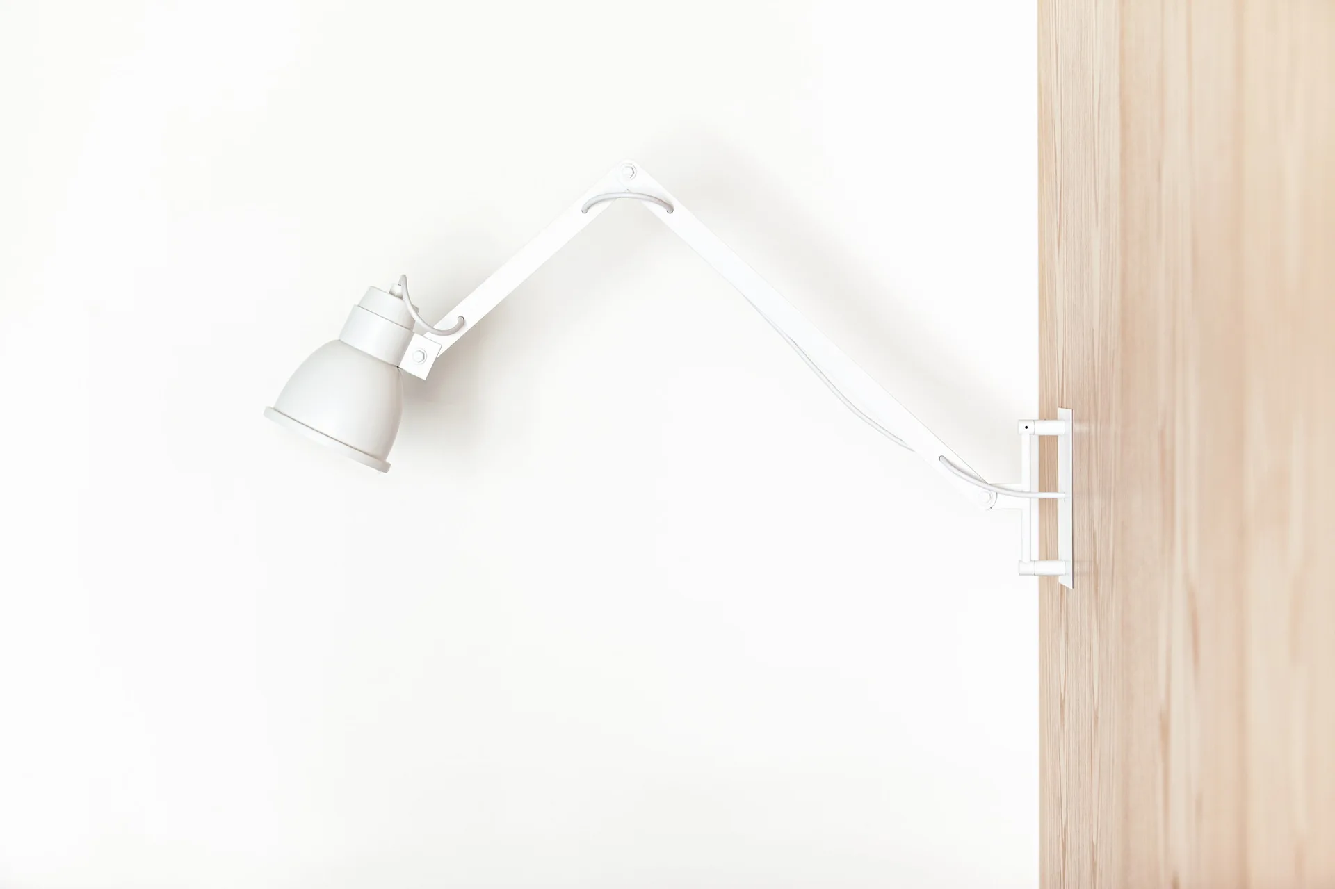 A matte white adjustable wall lamp with an articulated arm is mounted on a light wood panel next to a plain white wall.