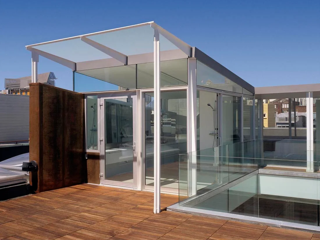 A modern rooftop terrace with a glass-enclosed shower structure, weathered steel partition, and warm wood plank decking.