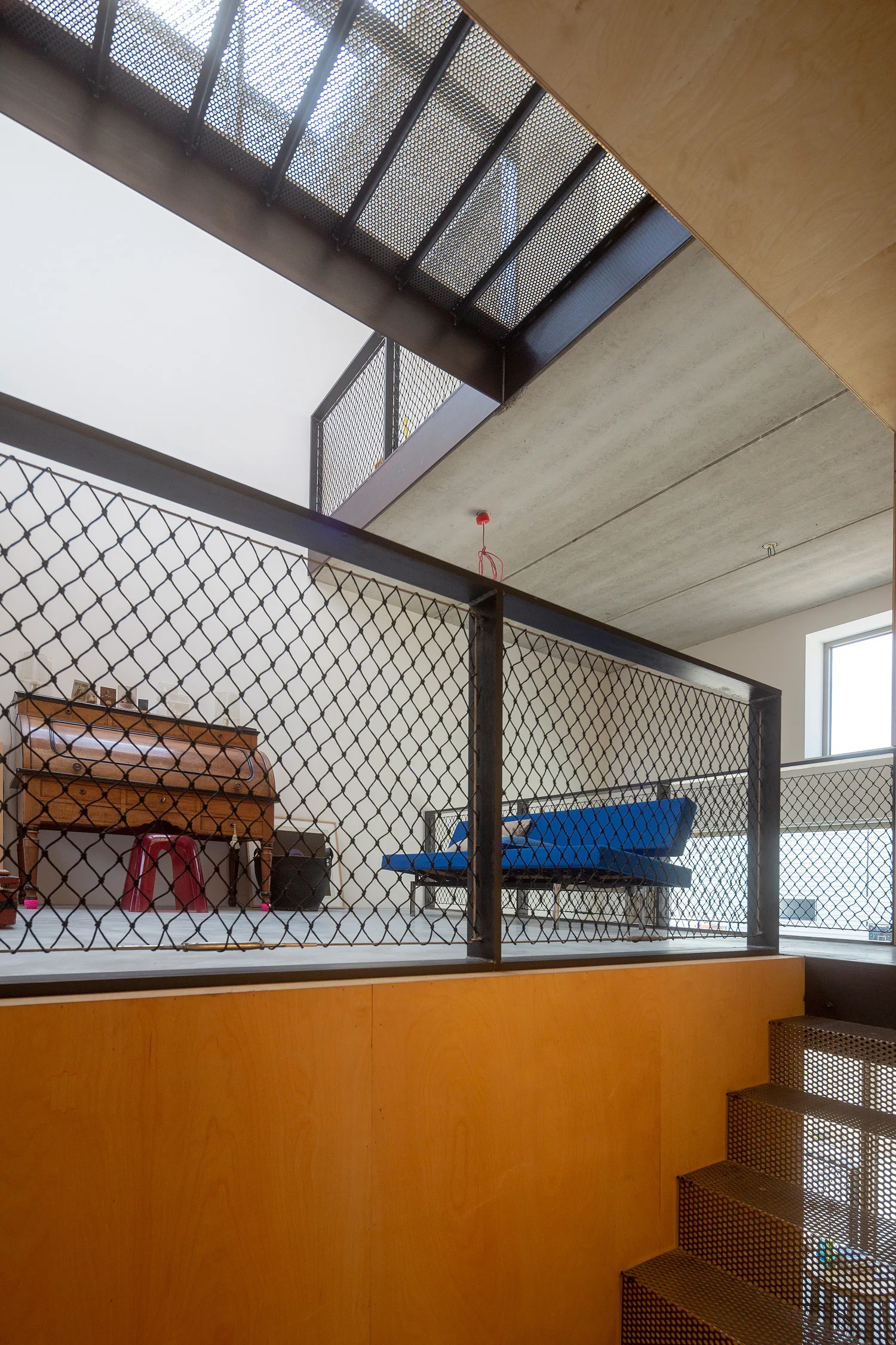 A modern loft mezzanine features black wire mesh safety railings, a bright blue sofa, a vintage wooden roll-top desk, and perforated metal stairs.