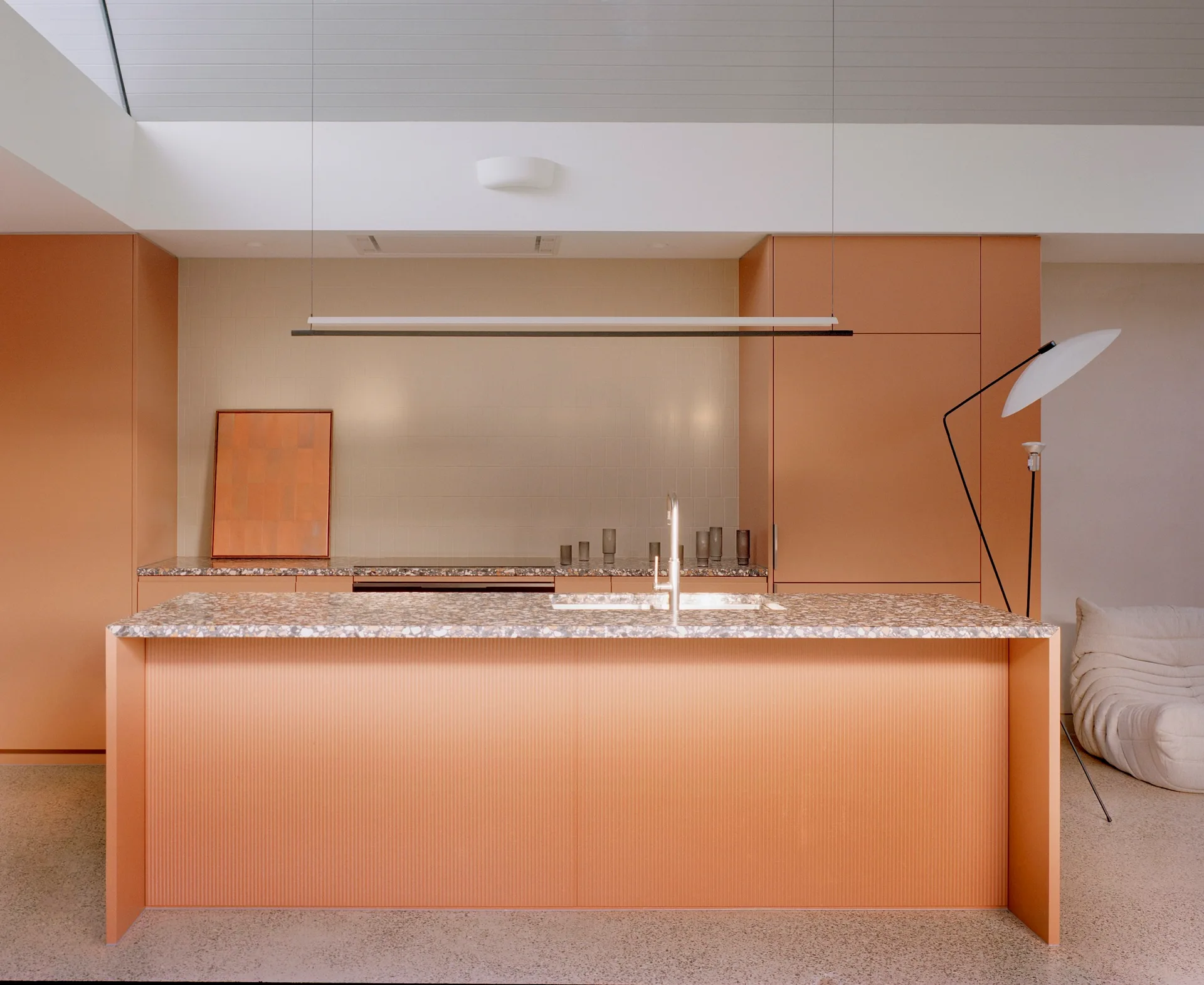 A modern kitchen featuring matte peach cabinetry, a ribbed kitchen island with a terrazzo countertop, and a simple cream tiled backsplash.