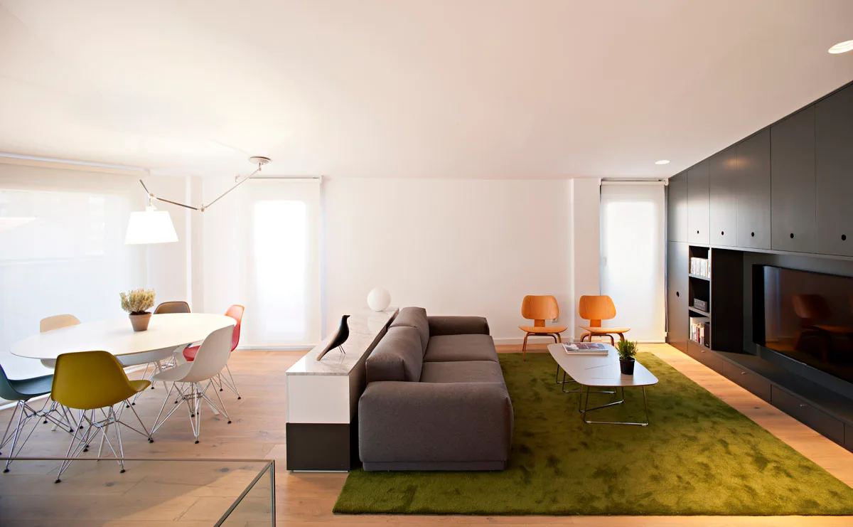 An open-plan living room features a grey sofa on a moss green rug, a matte black media wall, and an oval dining table with colorful chairs.