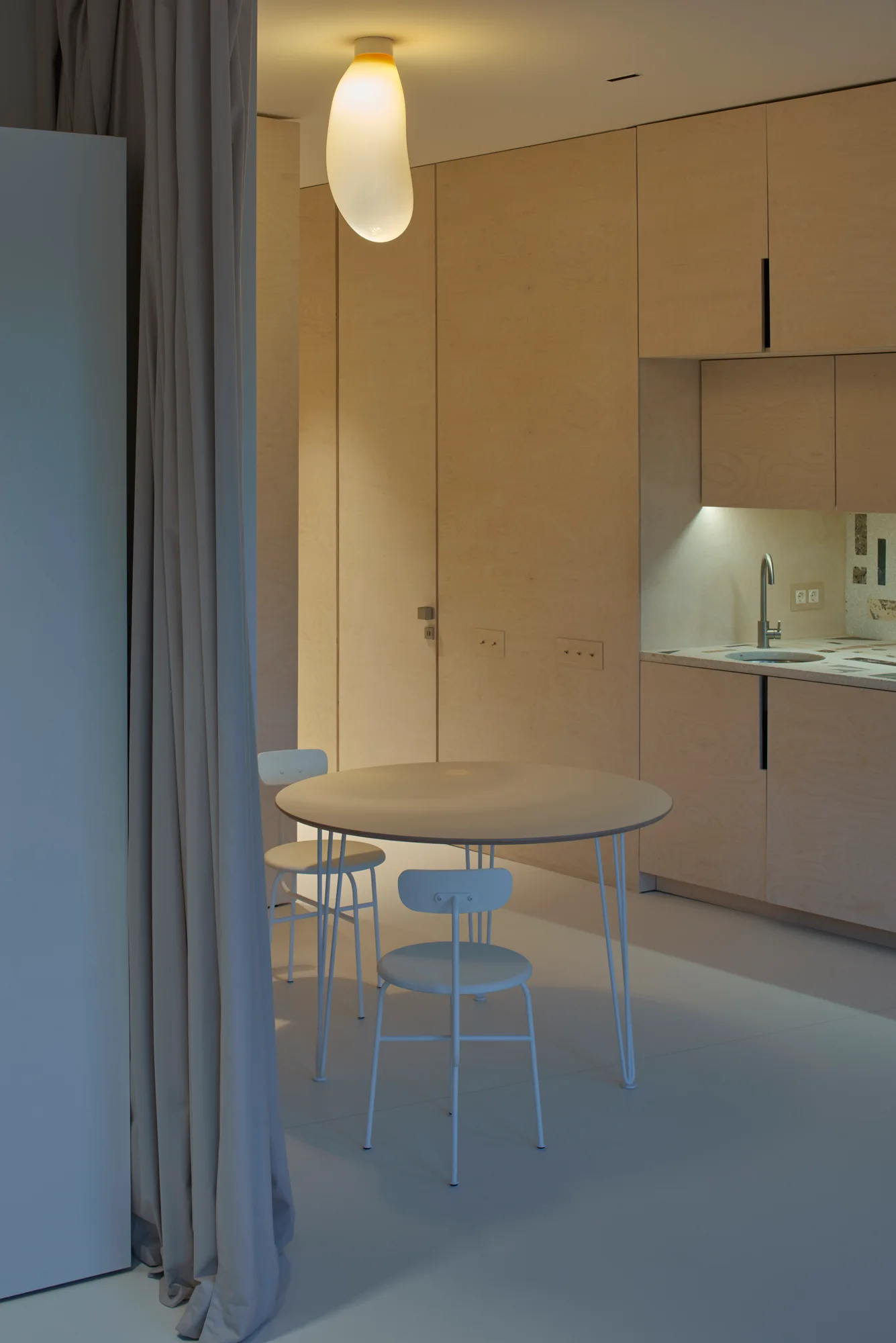 A round dining table and white chairs sit beside flat-front birch plywood kitchen cabinets under an organic-shaped glowing pendant light.