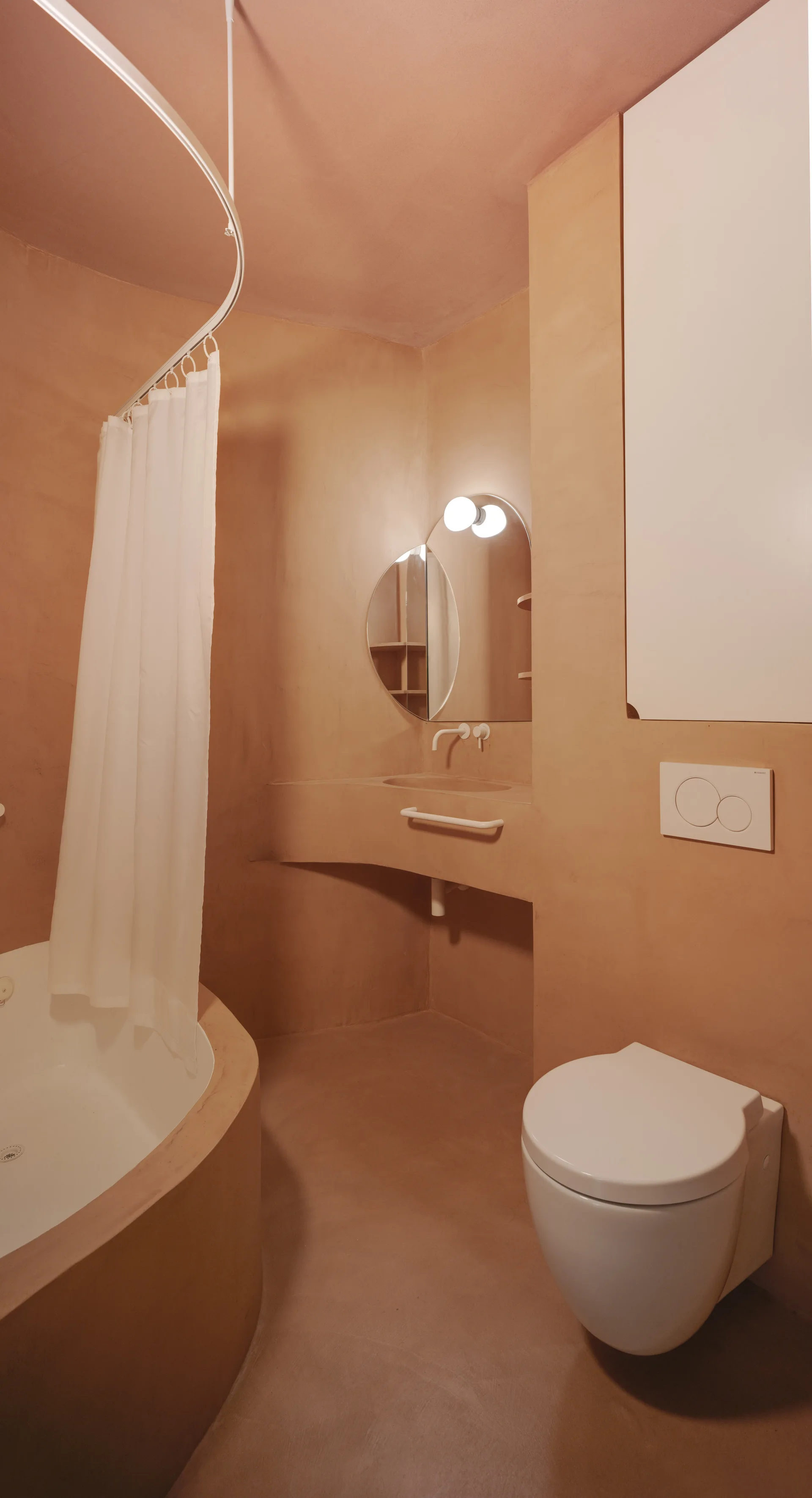 A minimalist bathroom features monochromatic terracotta plaster walls, a curved built-in vanity, a round mirror, and a wall-mounted toilet.