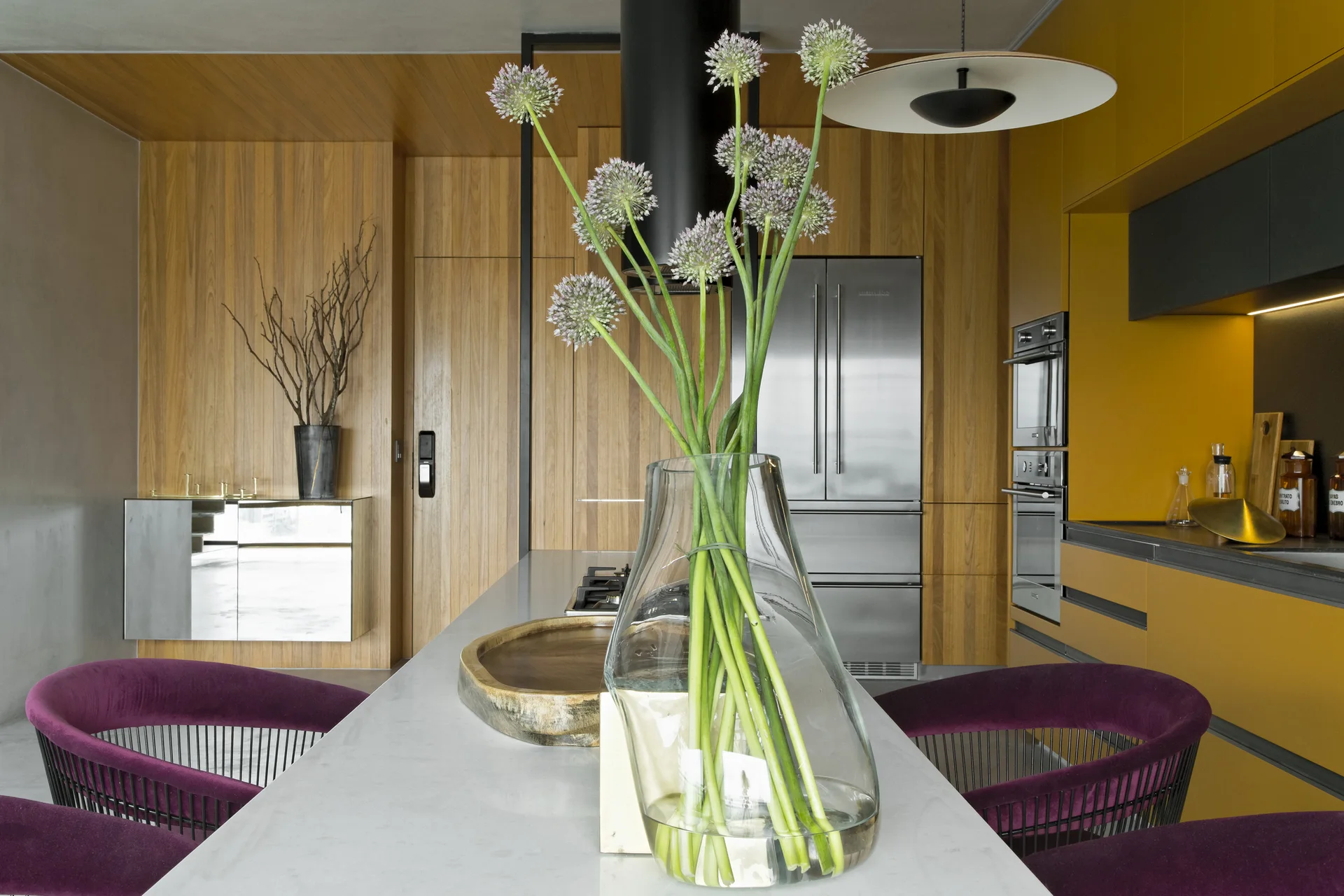 A modern kitchen features a stone island with purple armchairs, wood-paneled walls, a stainless steel refrigerator, and bold yellow cabinets.