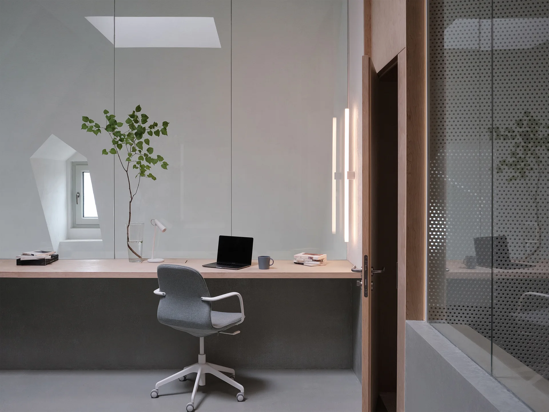 A minimalist office space featuring a long light wood floating desk, a grey swivel chair, and a clear glass partition wall.