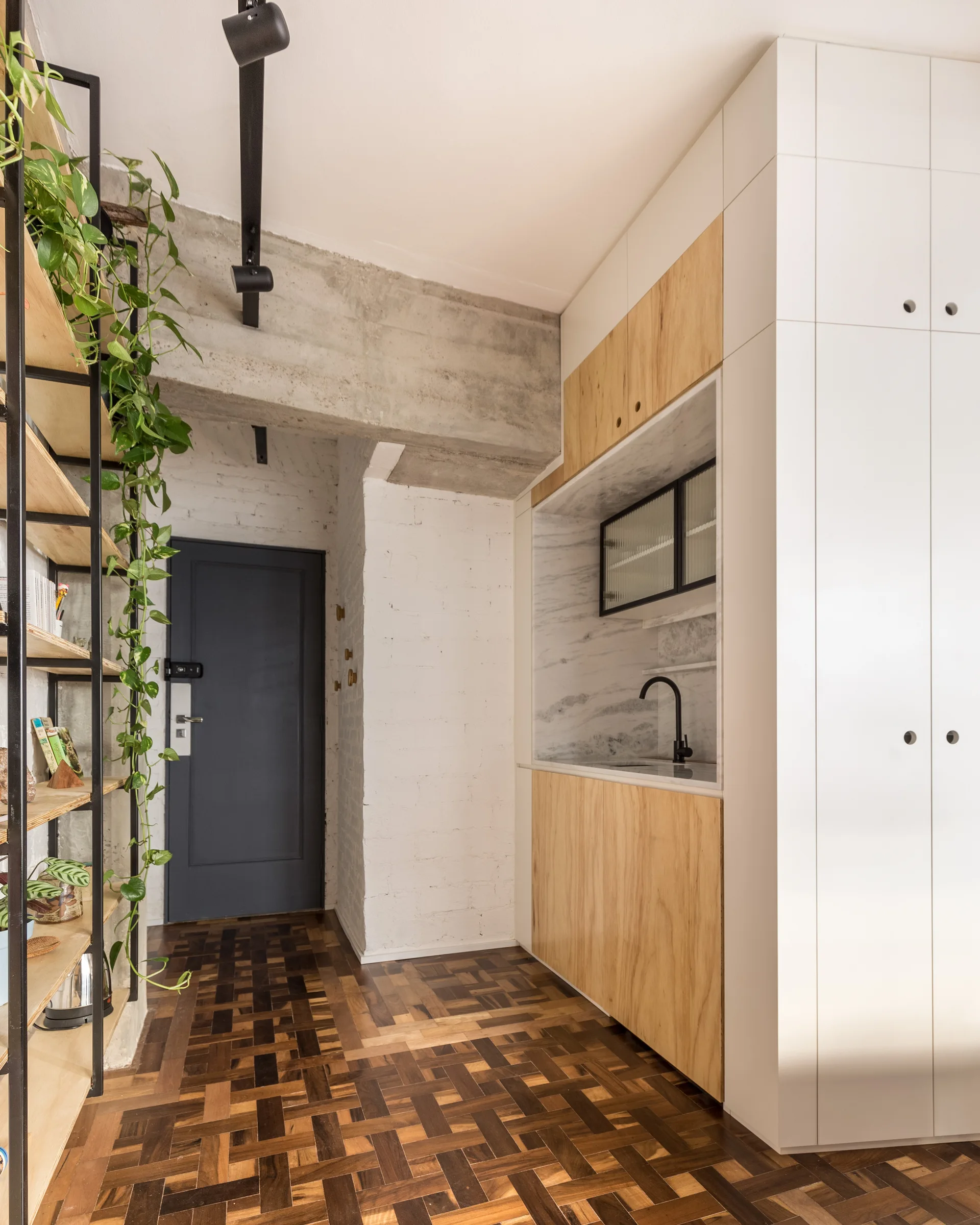 A residential entryway features a built-in kitchenette with wood cabinets, a dark patterned parquet floor, and black open metal shelving.