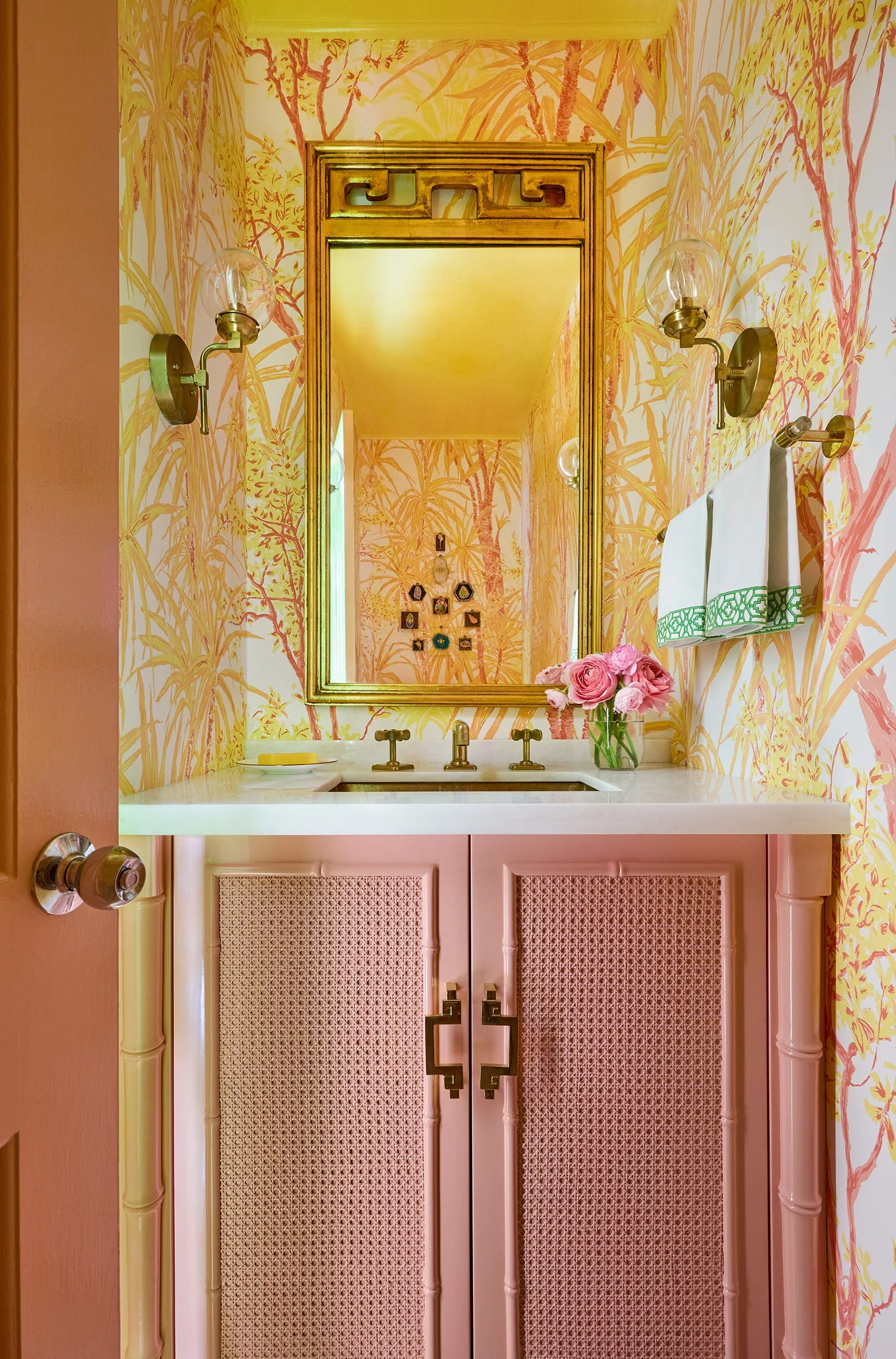 A powder room features a pink cane-front vanity, a gold-framed mirror, brass wall sconces, and vibrant yellow botanical wallpaper.