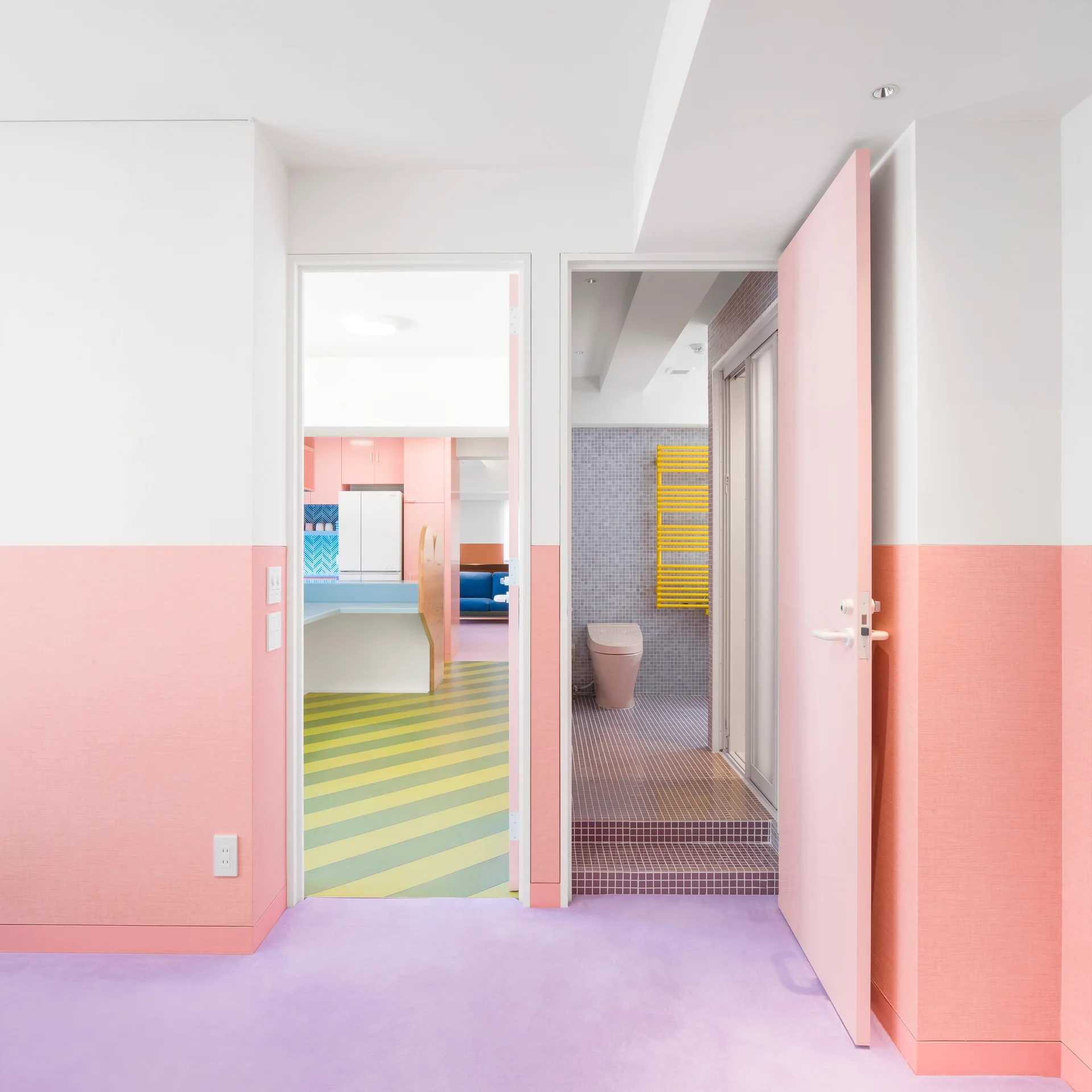 An interior hallway featuring pastel pink and white walls, lavender flooring, and doorways opening into a striped kitchen and a tiled bathroom.
