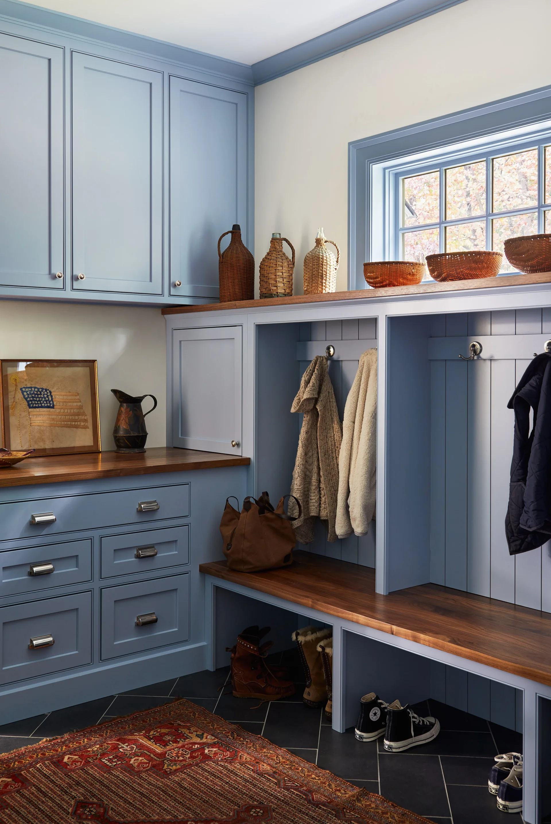 A mudroom featuring dusty blue built-in cabinets, a dark wood bench with coats, dark stone floor tiles, and a patterned rug.