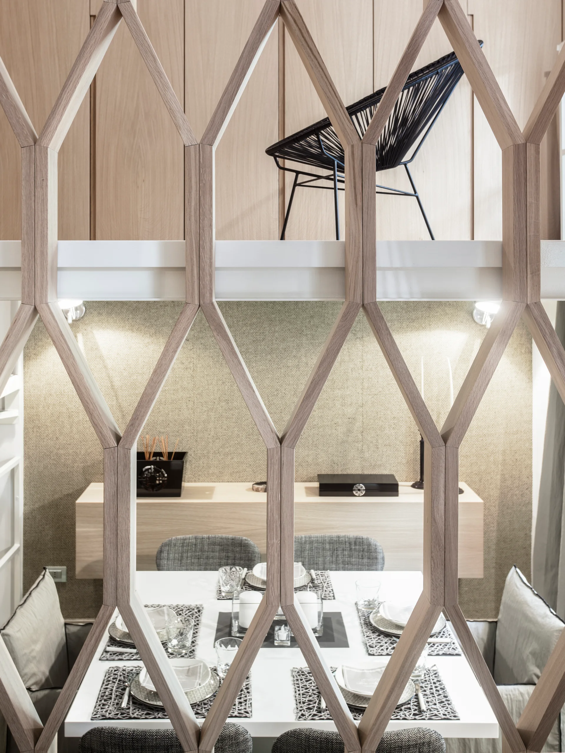 A geometric light wood screen partitions a dining area featuring a white table, woven placemats, and a floating shelf against a textured wall.