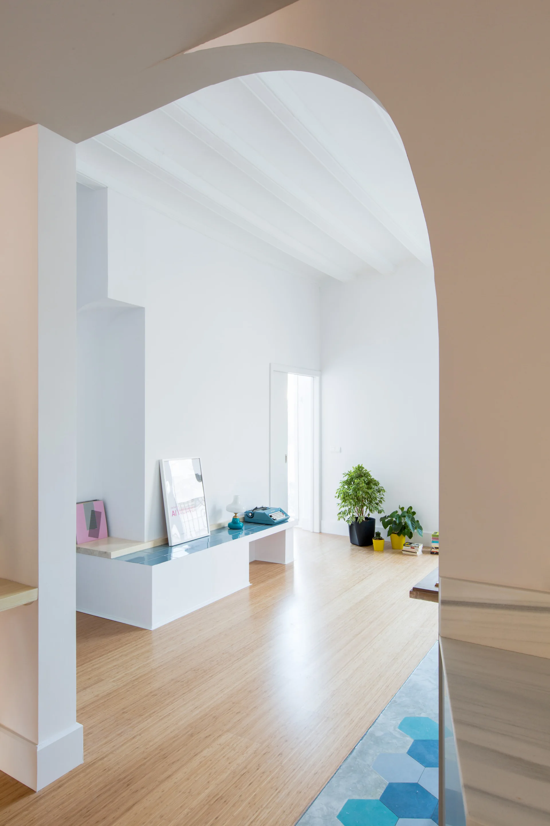 An arched doorway frames a minimalist white room with light wood floors, a built-in bench topped with blue tiles, and green potted plants.