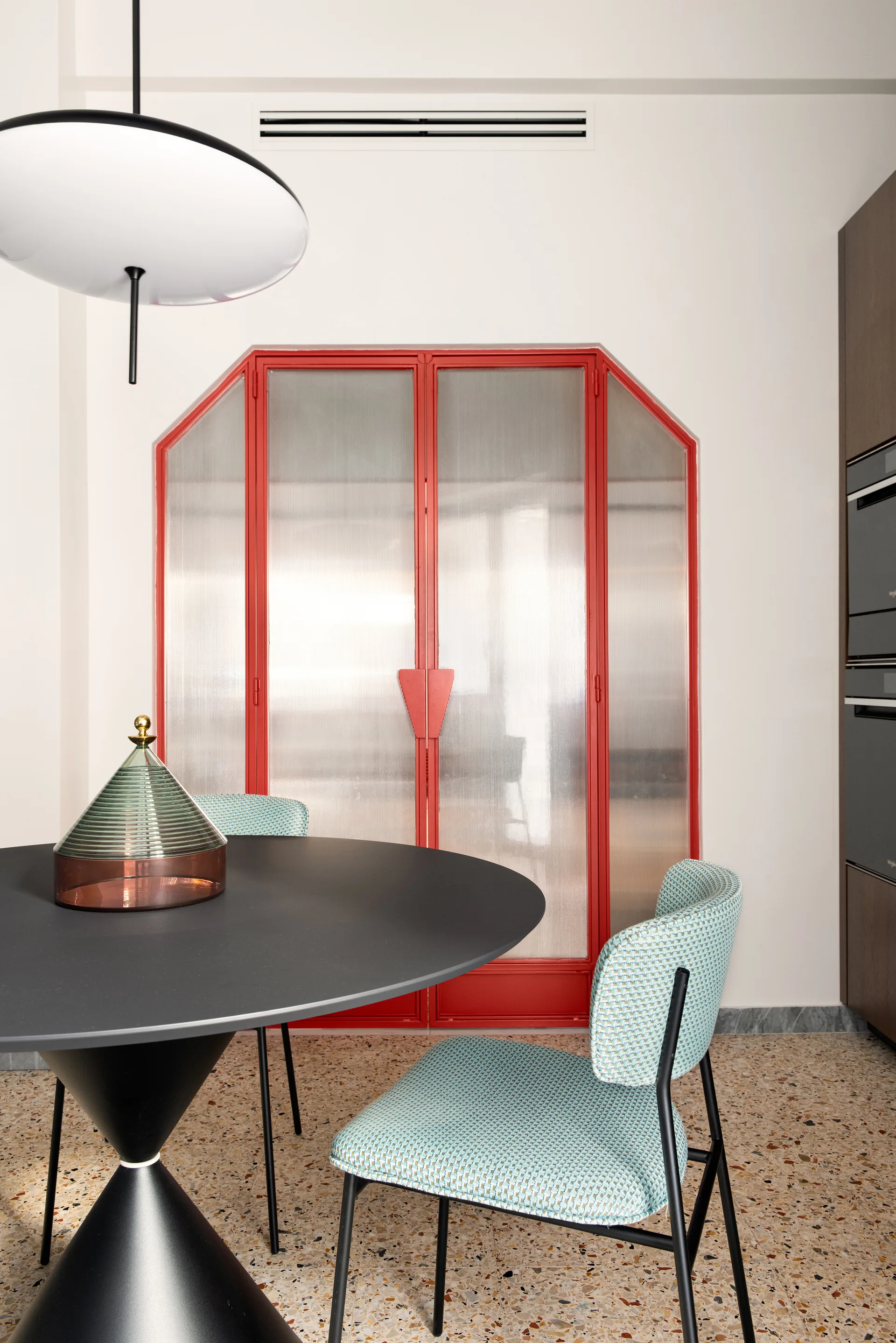A black circular dining table and turquoise patterned chairs stand on a terrazzo floor in front of red-framed reeded glass doors.