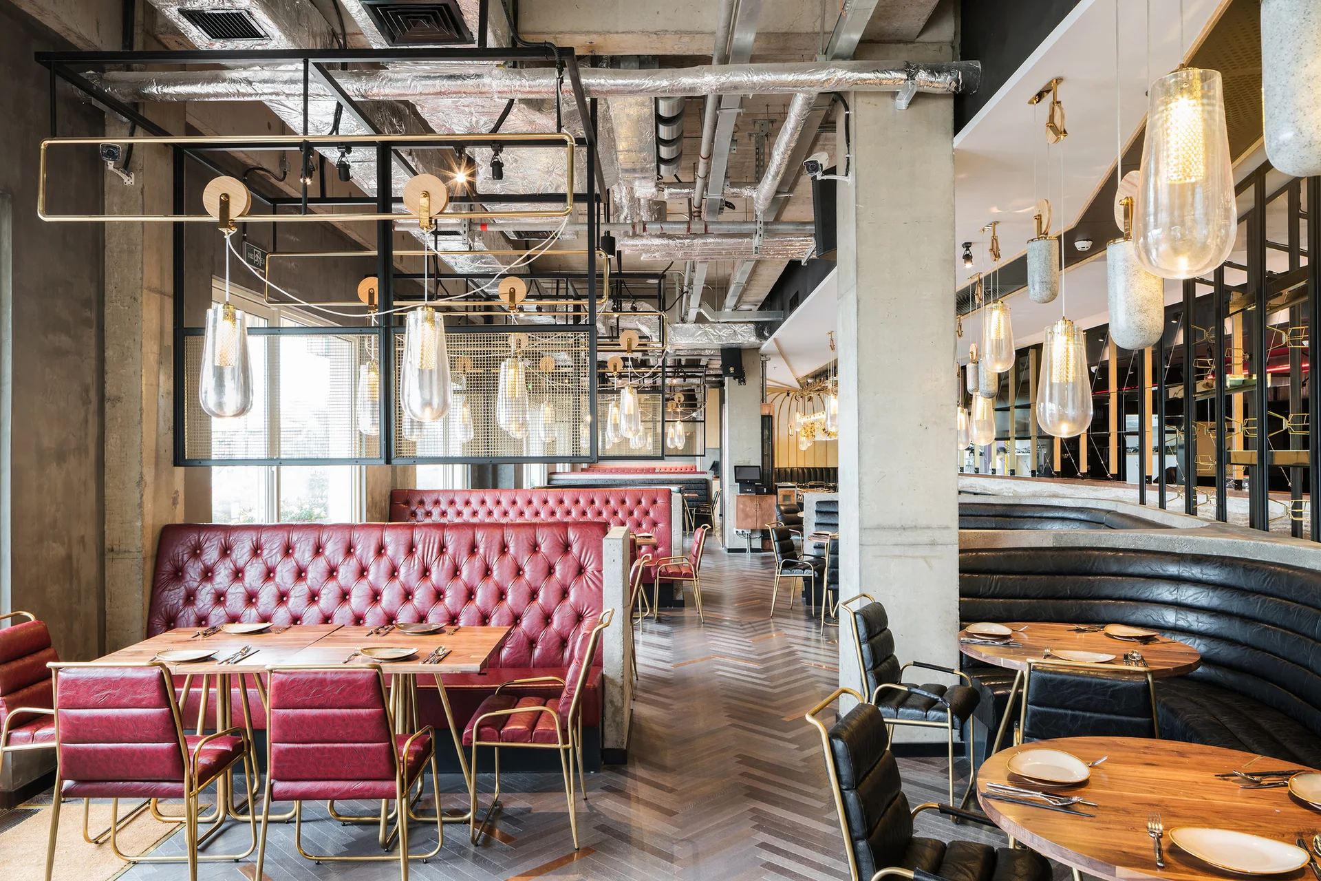 An industrial restaurant interior featuring tufted red leather booths, wooden tables, concrete columns, and exposed ceiling pipes overhead.
