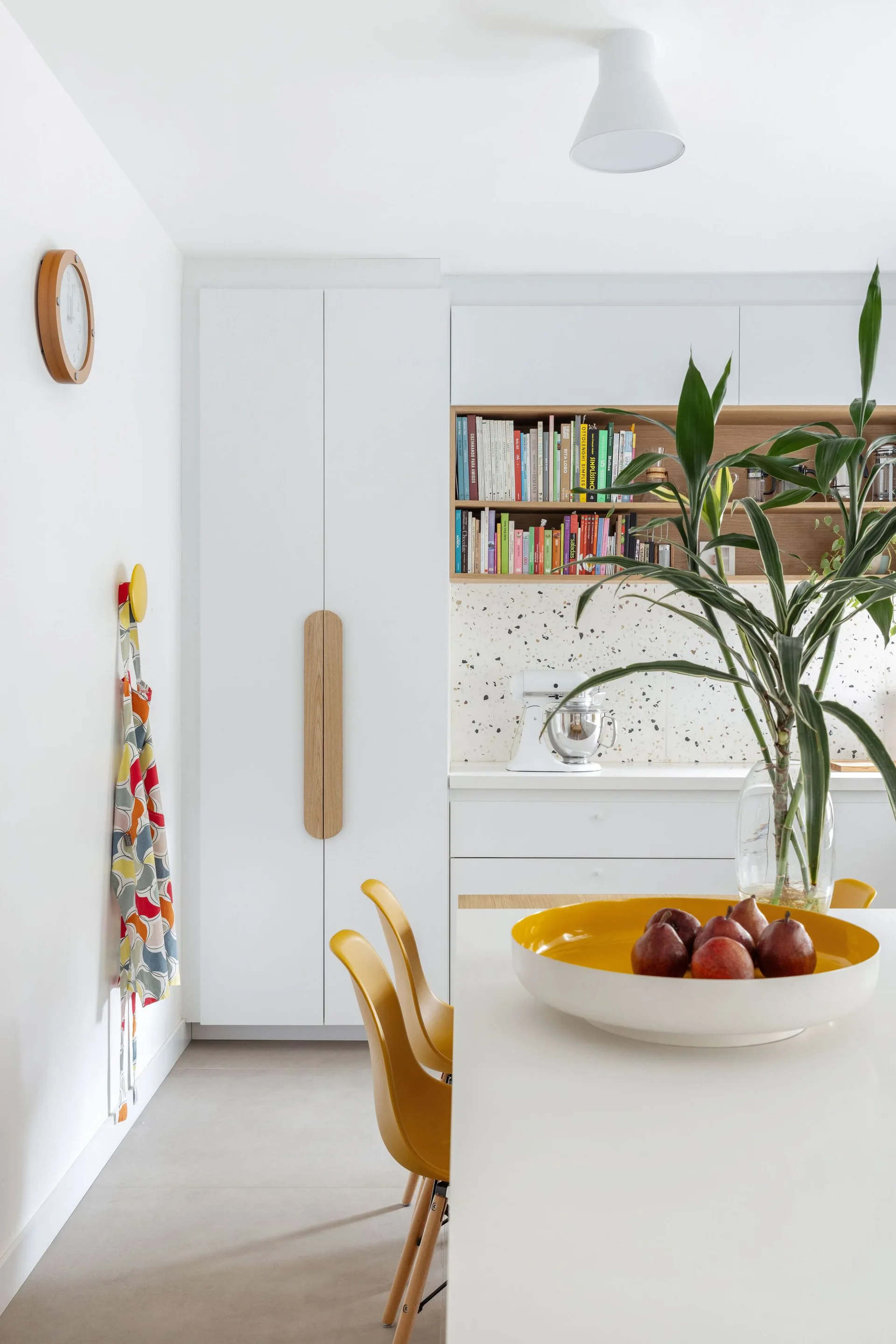 A modern white kitchen with wood-accented cabinets, a terrazzo backsplash, a built-in bookshelf, and yellow dining chairs.