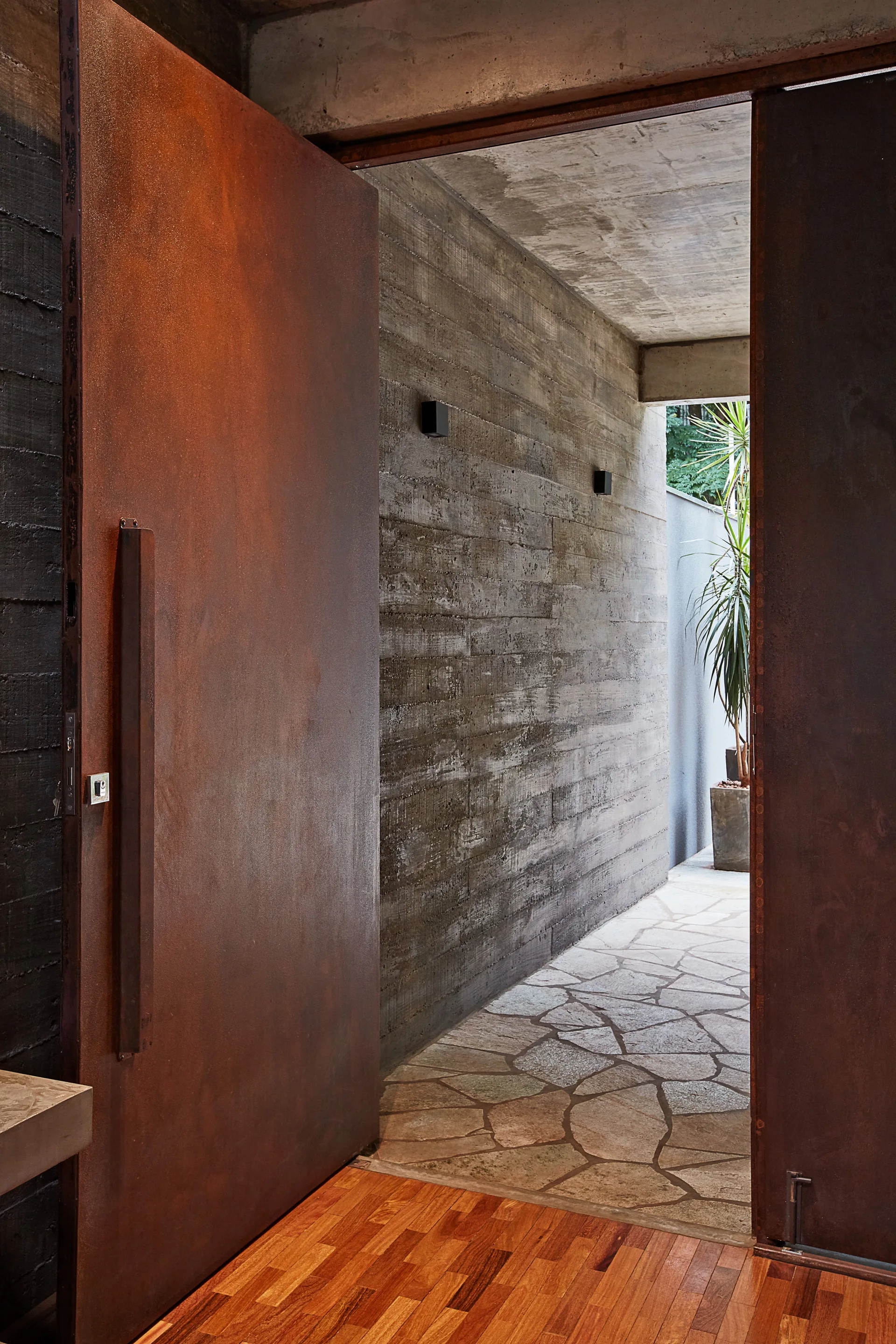 A large pivoting rusted metal door opens from a wood-floored entryway onto a flagstone path lined with board-formed concrete walls.