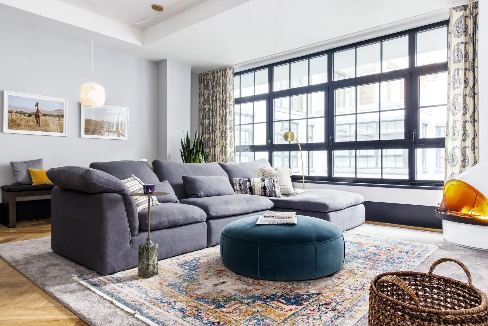 A grey sectional sofa and circular blue velvet ottoman sit on a distressed rug in front of large black-framed windows and a white retro fireplace.