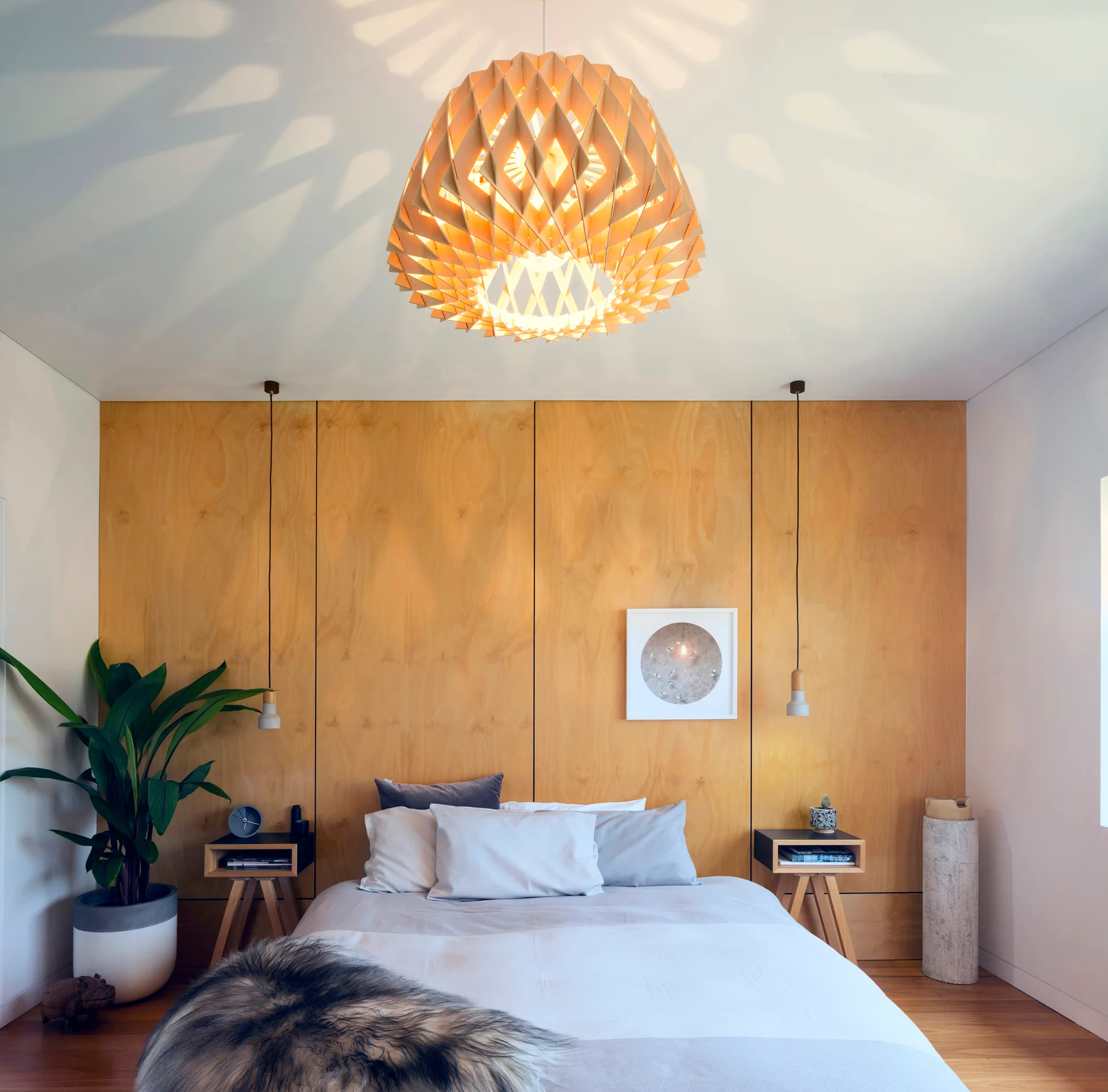A contemporary bedroom features a light wood paneled wall behind a bed with a large geometric wooden pendant light hanging above.