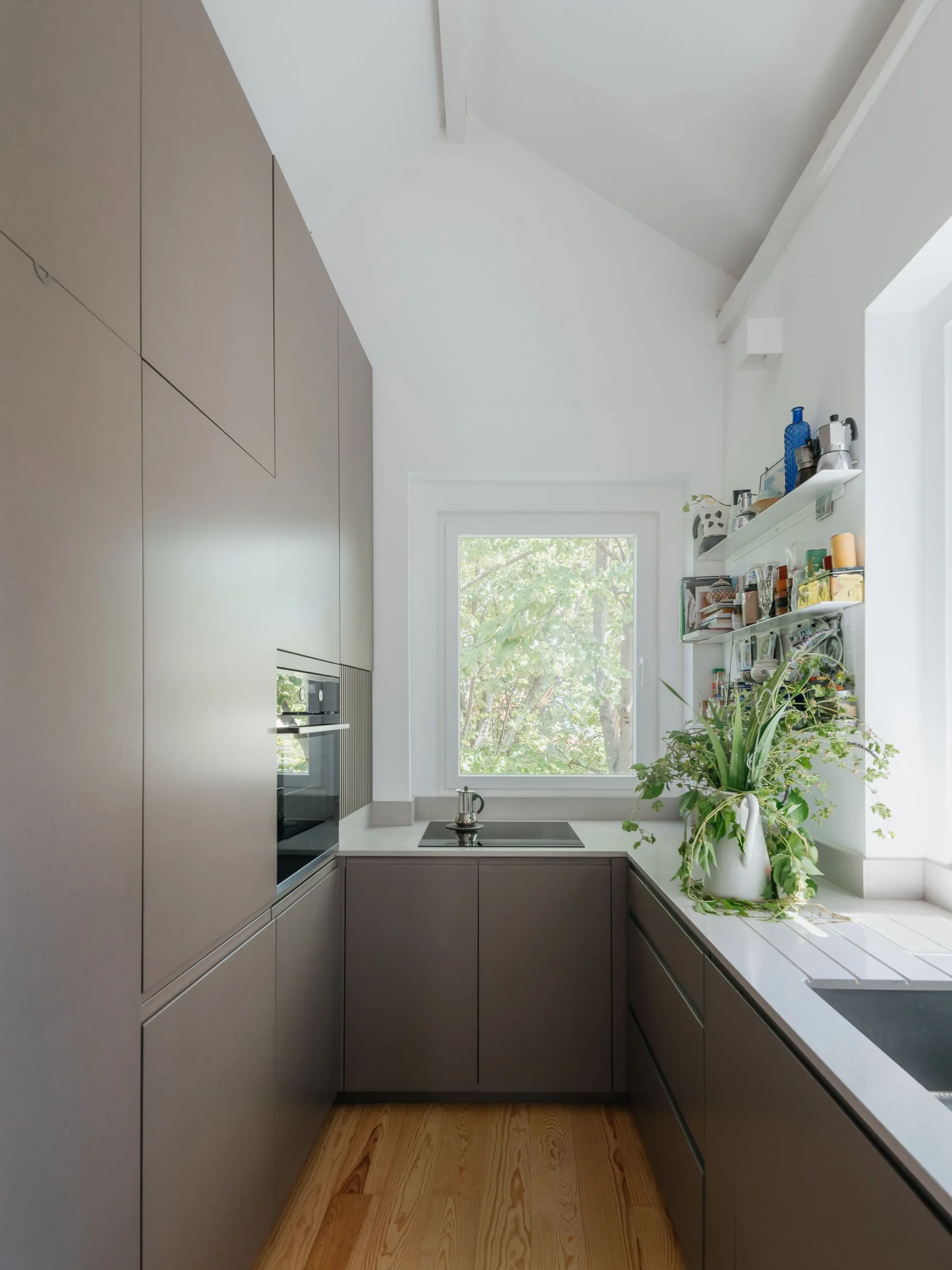 A compact minimalist kitchen with matte taupe flat-panel cabinets, light wood flooring, and a window framed by green trees.