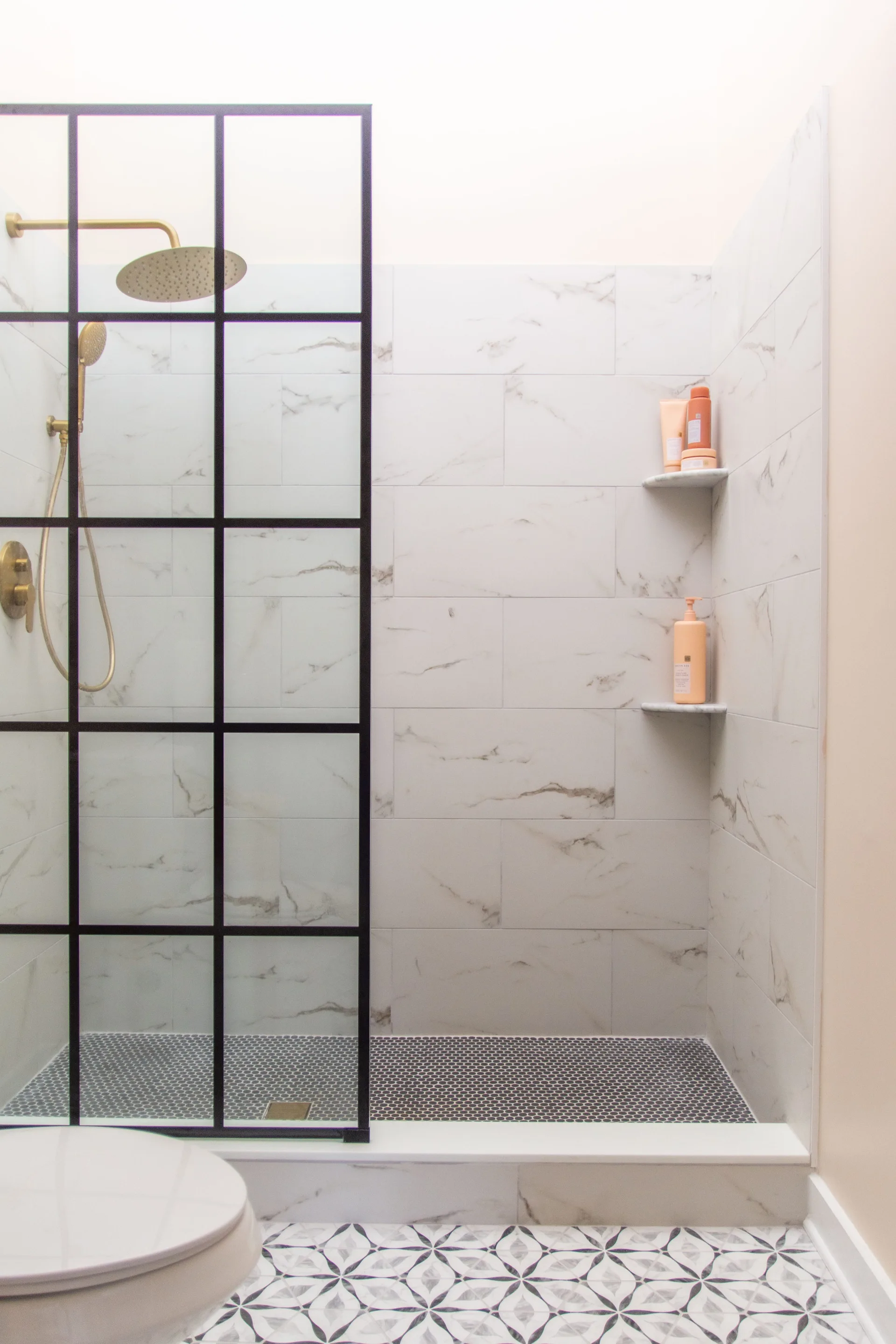 A modern walk-in shower features large marble-patterned wall tiles, a black metal grid-frame glass partition, brass fixtures, and dark penny tile flooring.