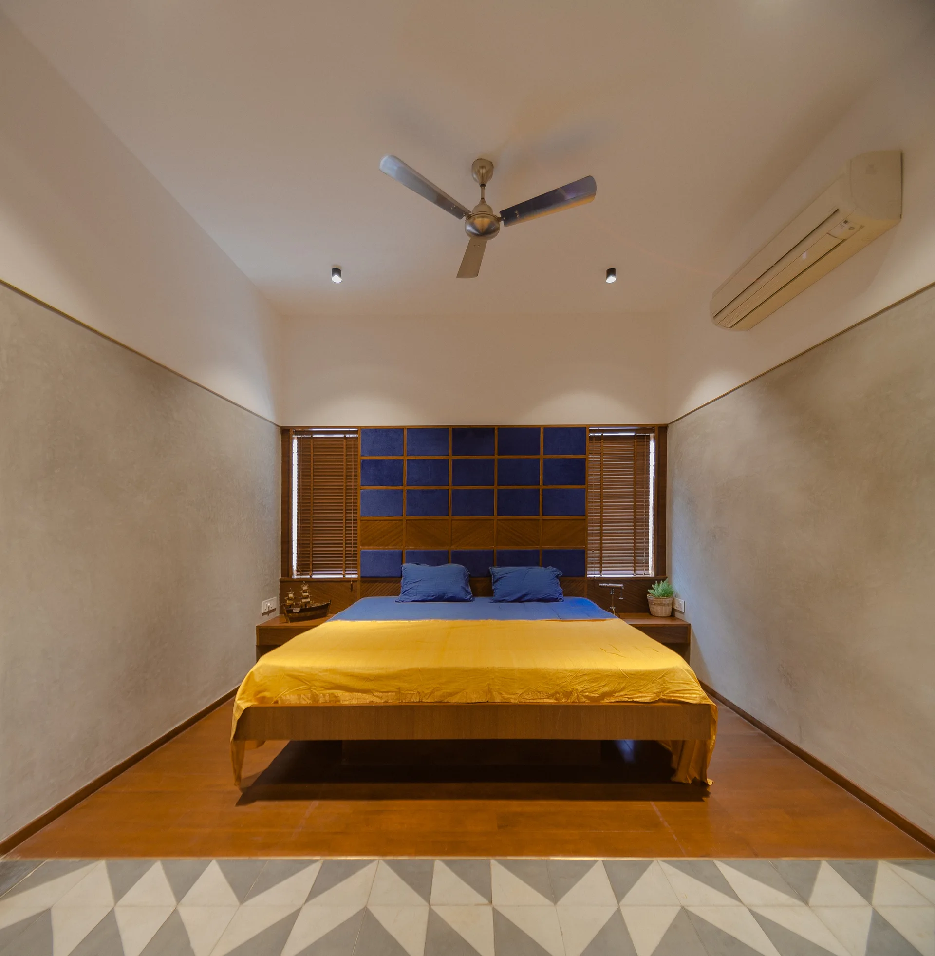 A modern bedroom features a bed with yellow bedding, a blue grid-patterned upholstered headboard, wooden blinds, and textured gray walls.