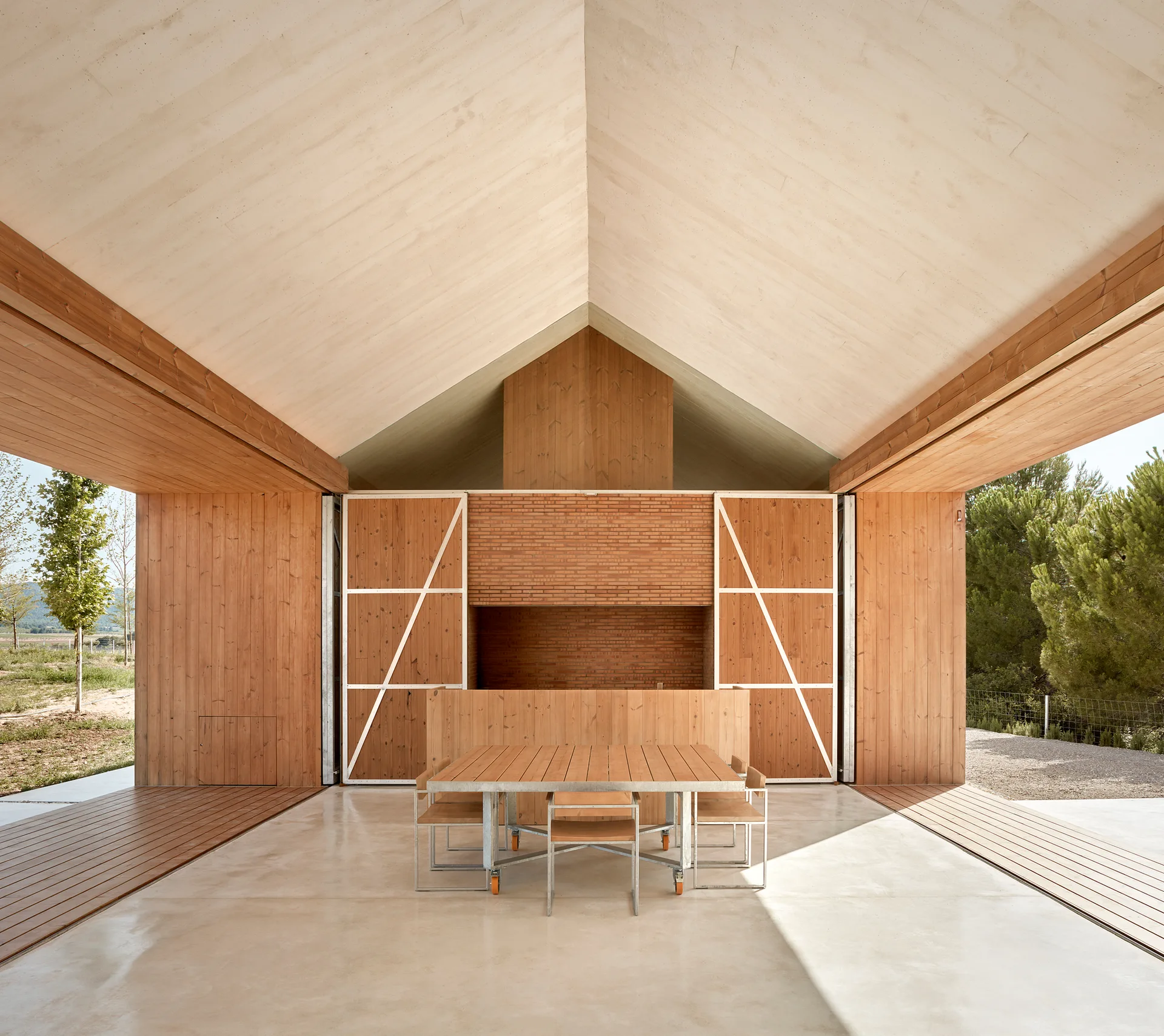 A covered outdoor patio features a gabled wood ceiling, timber-clad walls, a brick fireplace, and a wooden dining set on a smooth concrete floor.