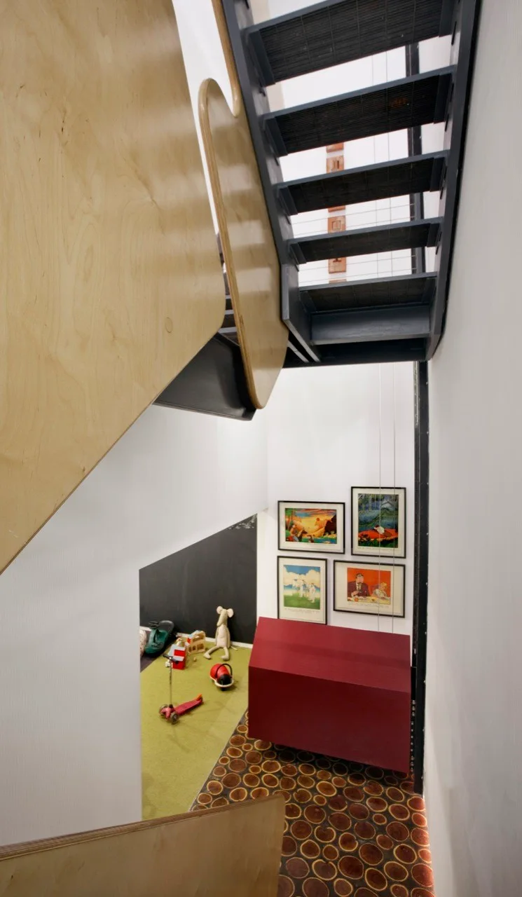 A high-angle view looking down a modern metal staircase with light curved plywood railings onto a landing with wood slice flooring and a red cabinet.