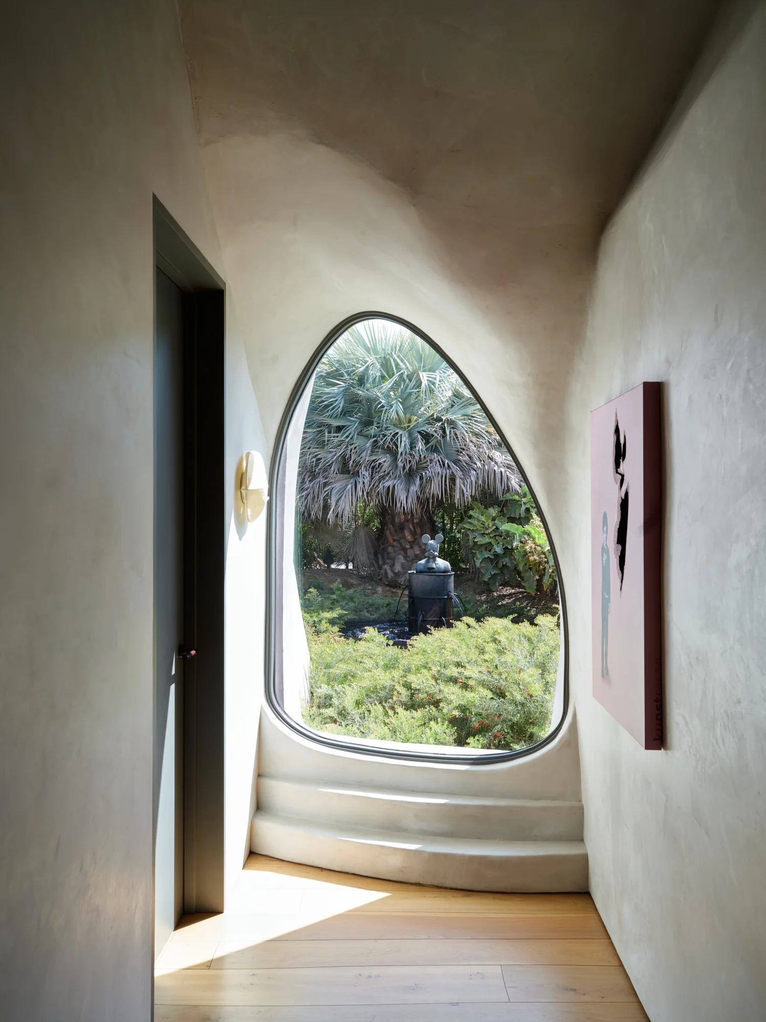 A minimalist hallway with smooth plaster walls, wood flooring, and an organic asymmetrical window framing a lush garden view.