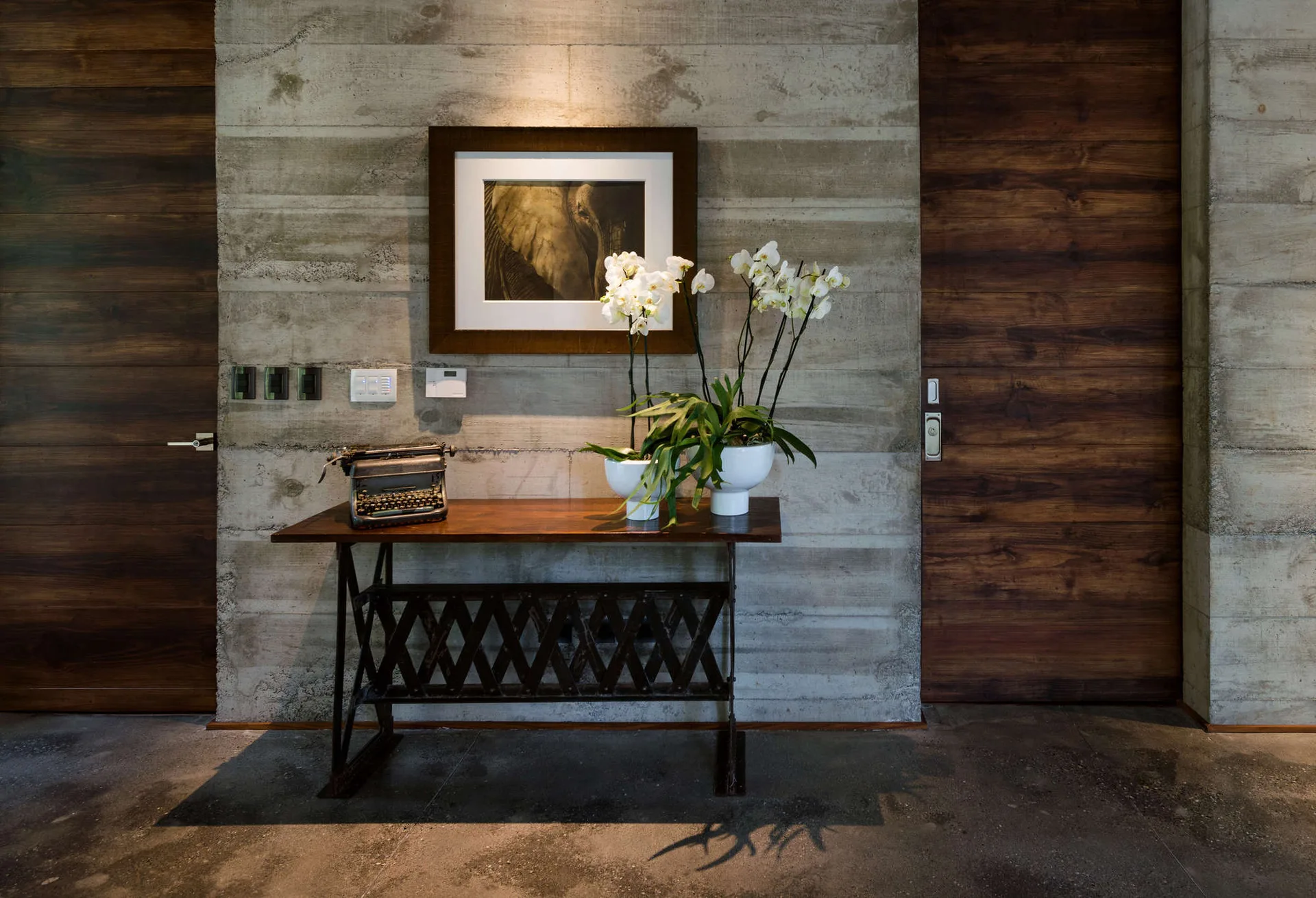 A vintage typewriter and potted white orchids sit on a wood console table against a board-formed concrete wall flanked by dark wood doors.