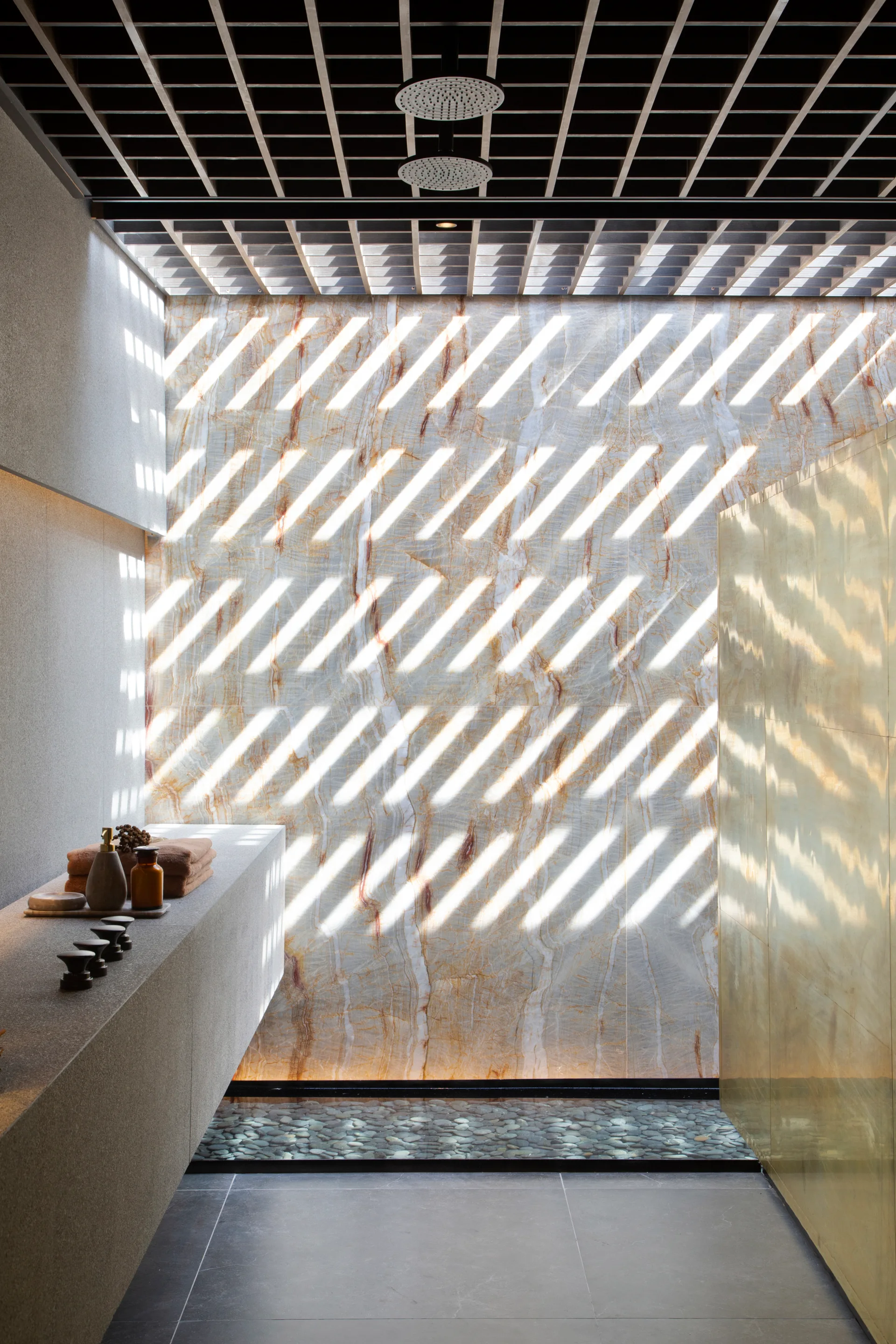 A modern shower with a highly veined stone wall, a black ceiling grid casting striped shadows, a stone ledge, and a pebble drain.