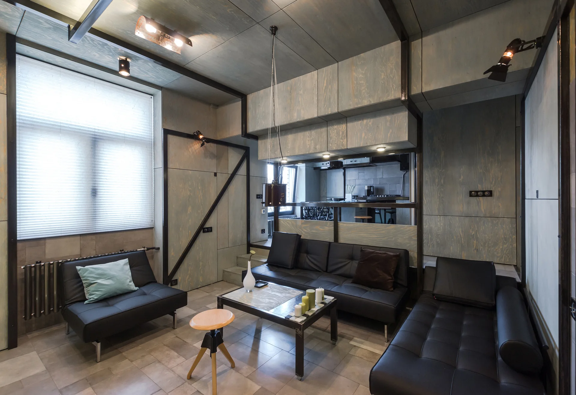 A modern living room features blue-grey stained plywood wall paneling, black metal framing, and low-profile black leather sofas on a grey tile floor.