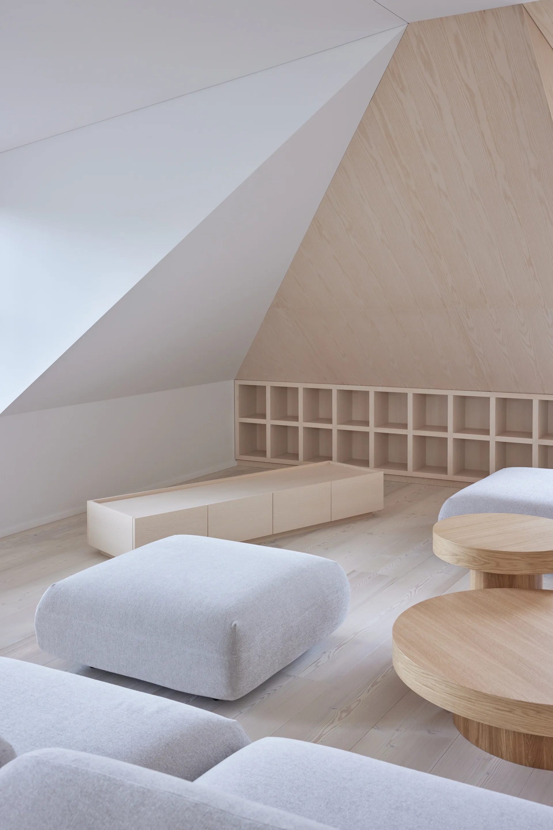 A minimalist attic lounge features a low gray sofa, round wooden coffee tables, and built-in grid shelving beneath a sloped wooden ceiling.