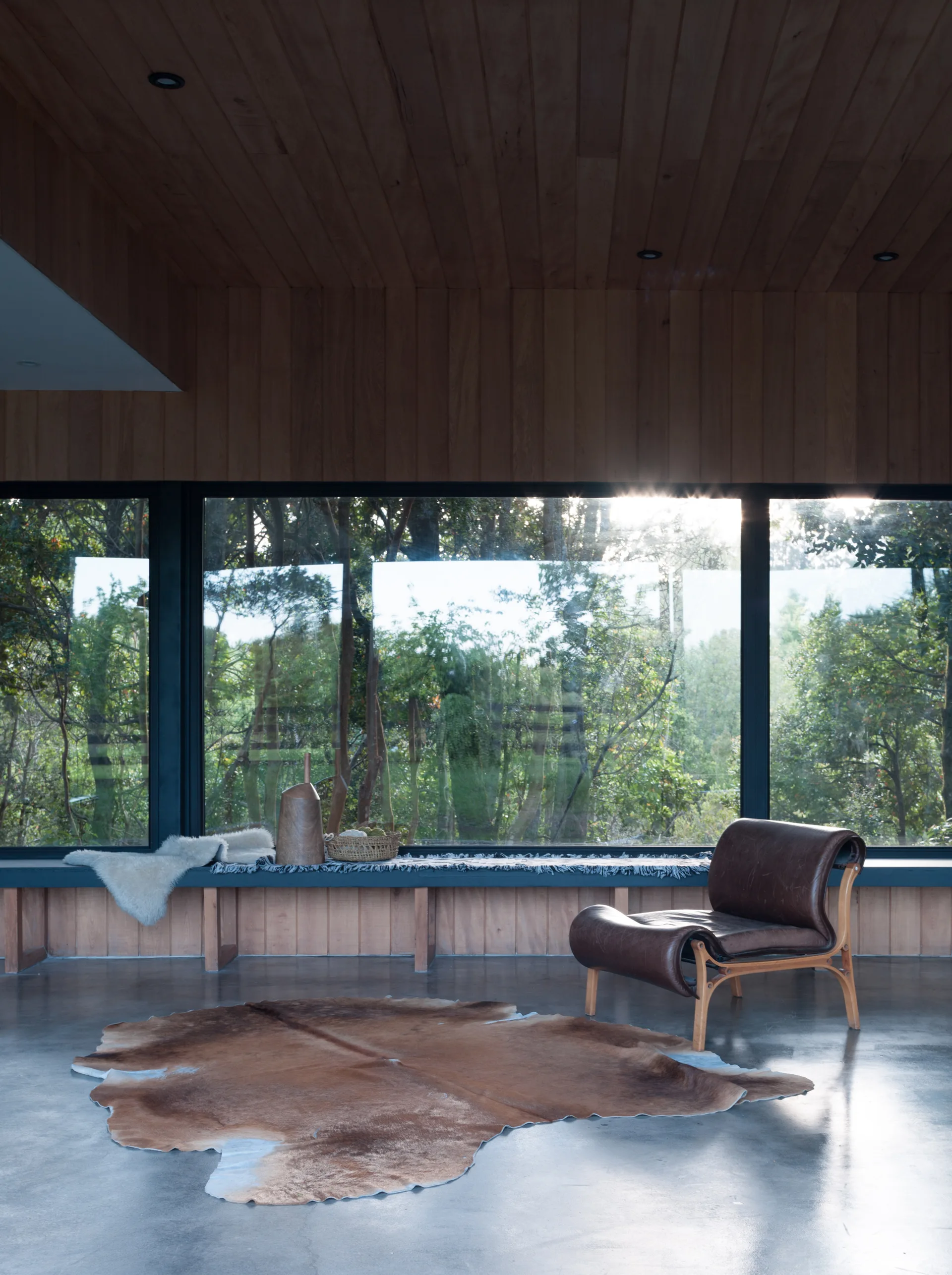 A modern room features a brown leather lounge chair on a cowhide rug in front of a large window looking onto a dense green forest.