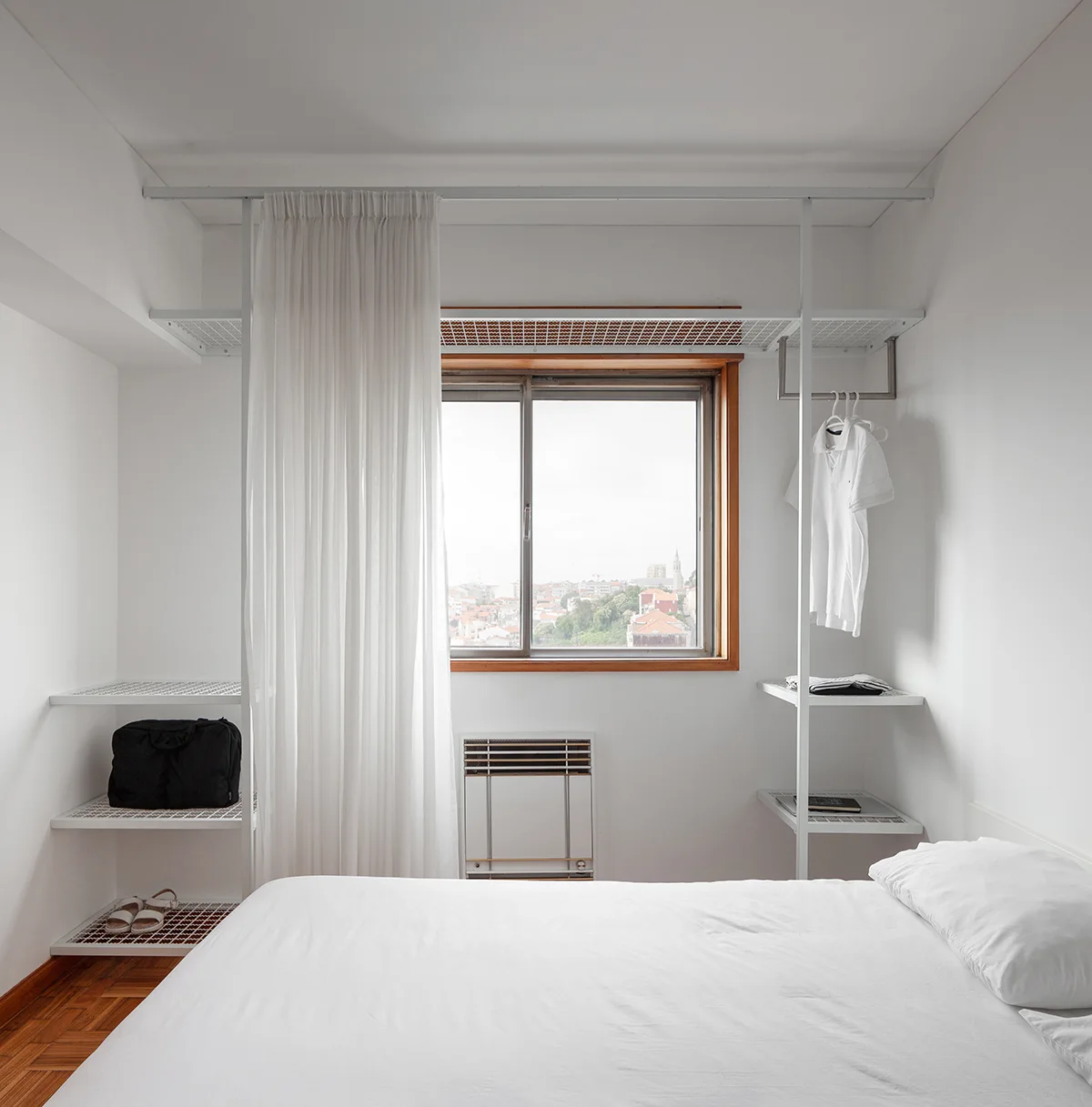 A minimalist white bedroom features a bed with white linens and an open metal closet system surrounding a wood-trimmed window.